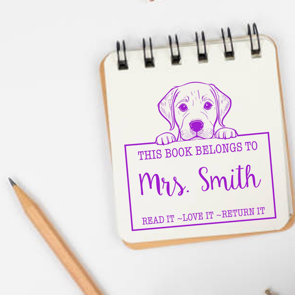 Purple custom book stamp with dog and This Book Belongs to Mrs. Smith on a white notepad, pencil beside it.