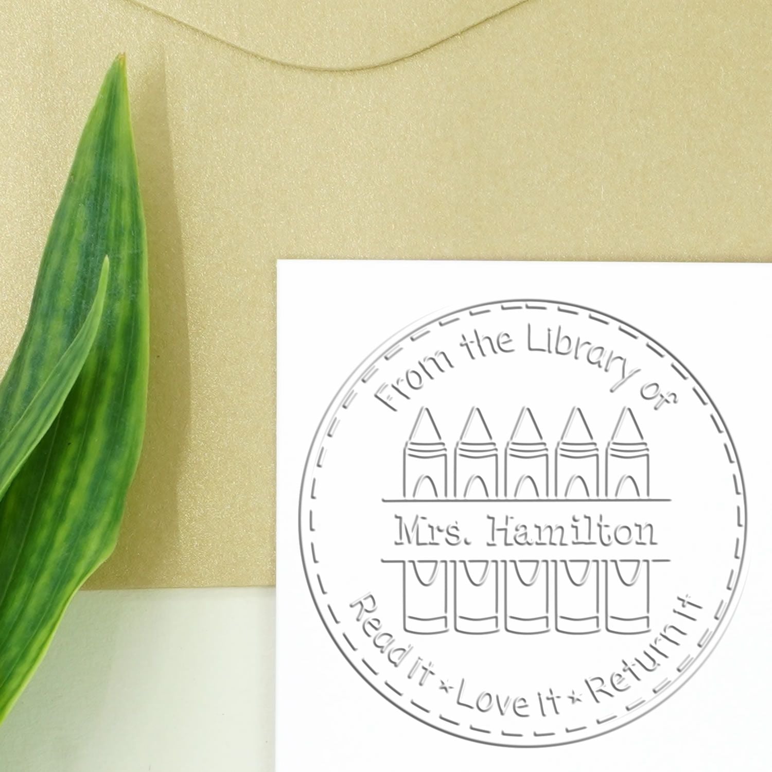 White circular library stamp seal with gray pencils and text From the Library of Mrs. Hamilton; green leaf beside it.