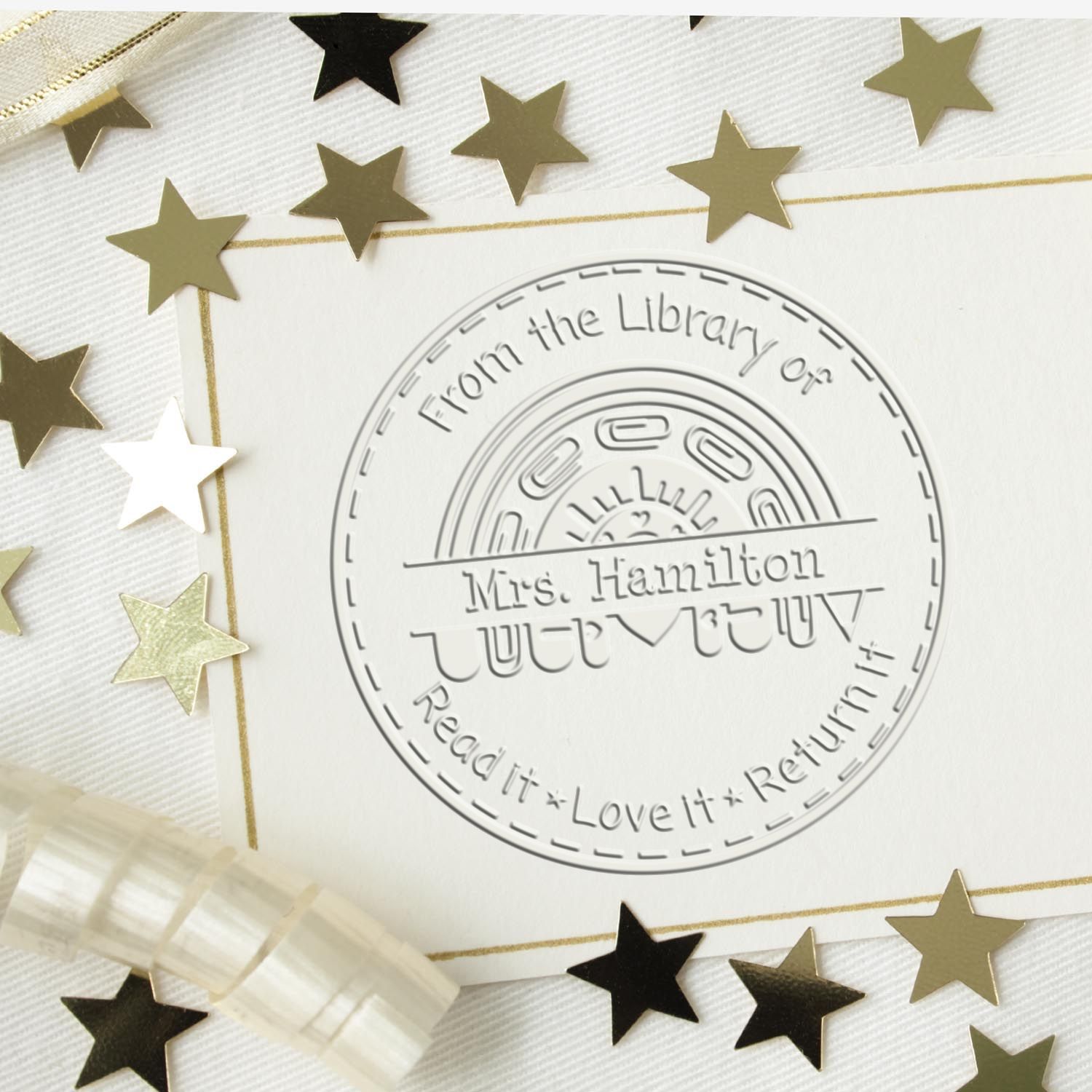 Embossed round library stamp and seal on white card, with gold and black stars and gold trim, reading Mrs. Hamilton.