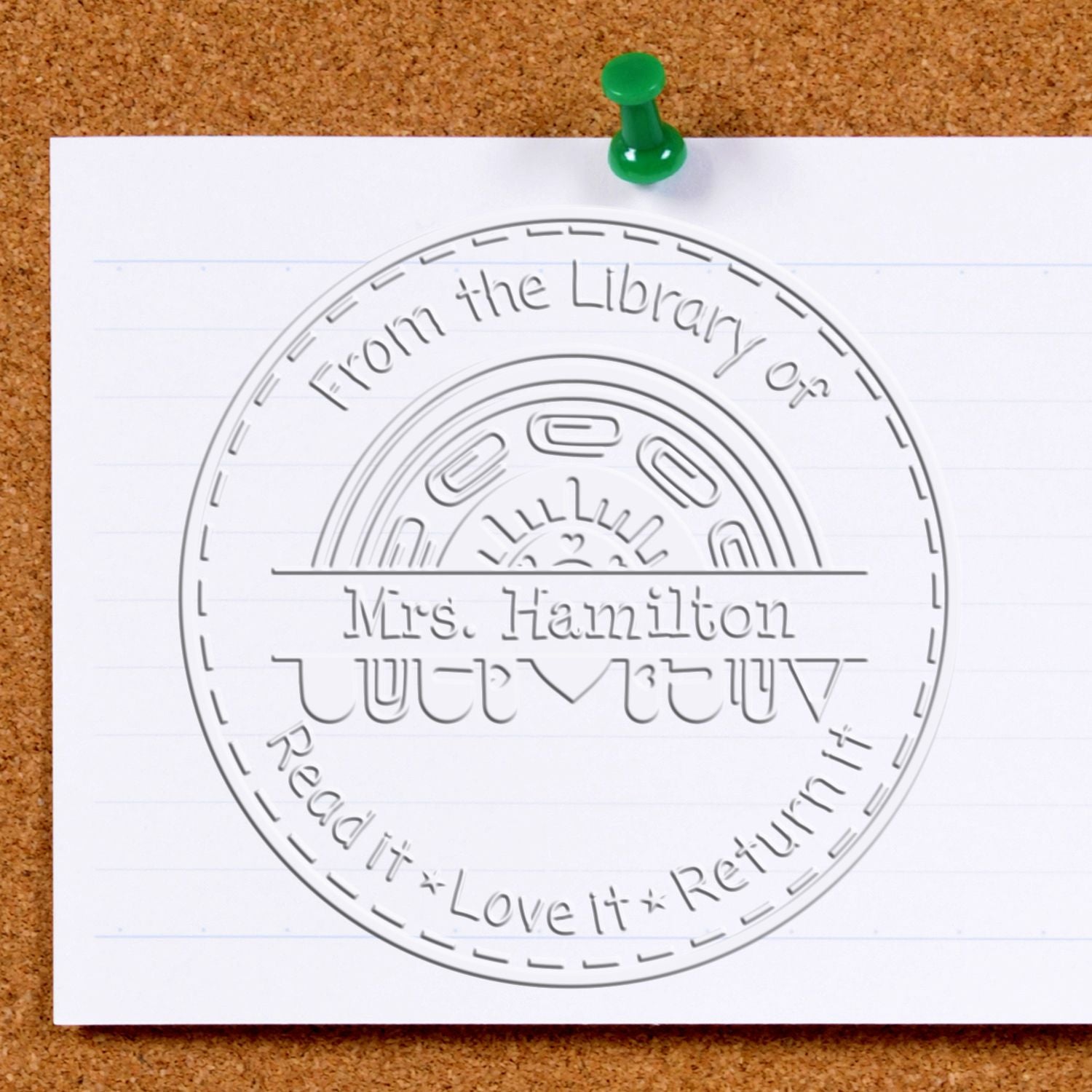 Green pushpin on white paper showing an embossed round library return stamp seal with Mrs. Hamilton on a cork board.