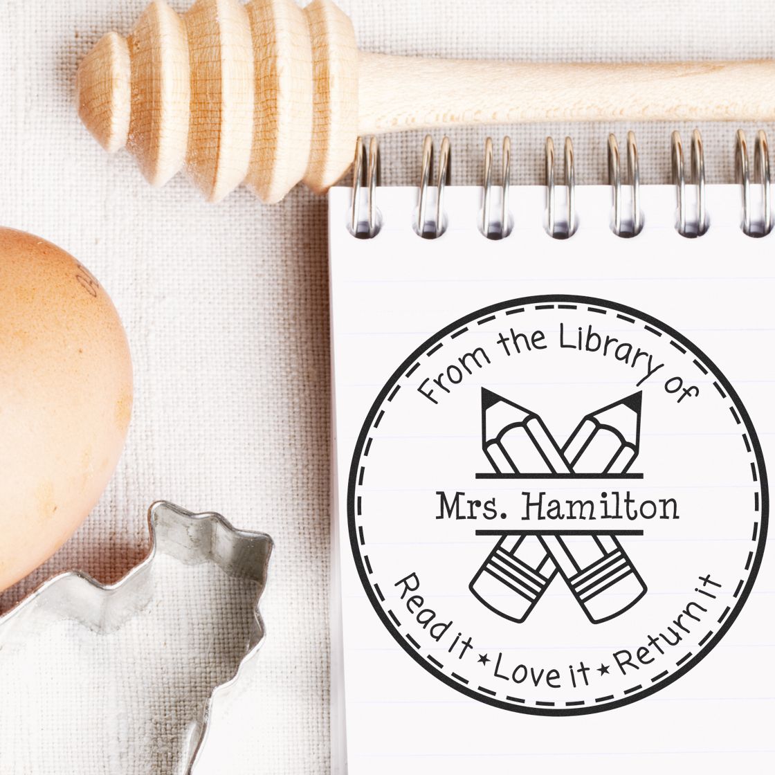 Black custom round library stamp seal with pencil icon and Mrs. Hamilton on a white spiral notebook beside a wooden stamp.