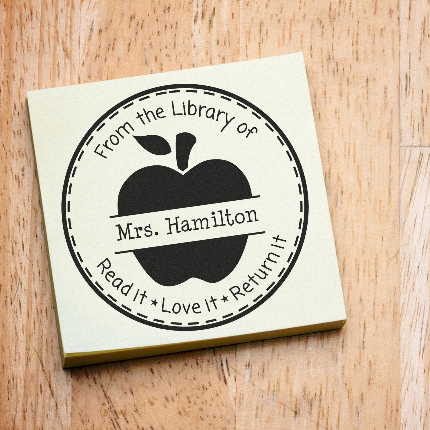 Black custom stamp and seal for library returns, with apple icon, Mrs. Hamilton, on a cream block over wood grain.