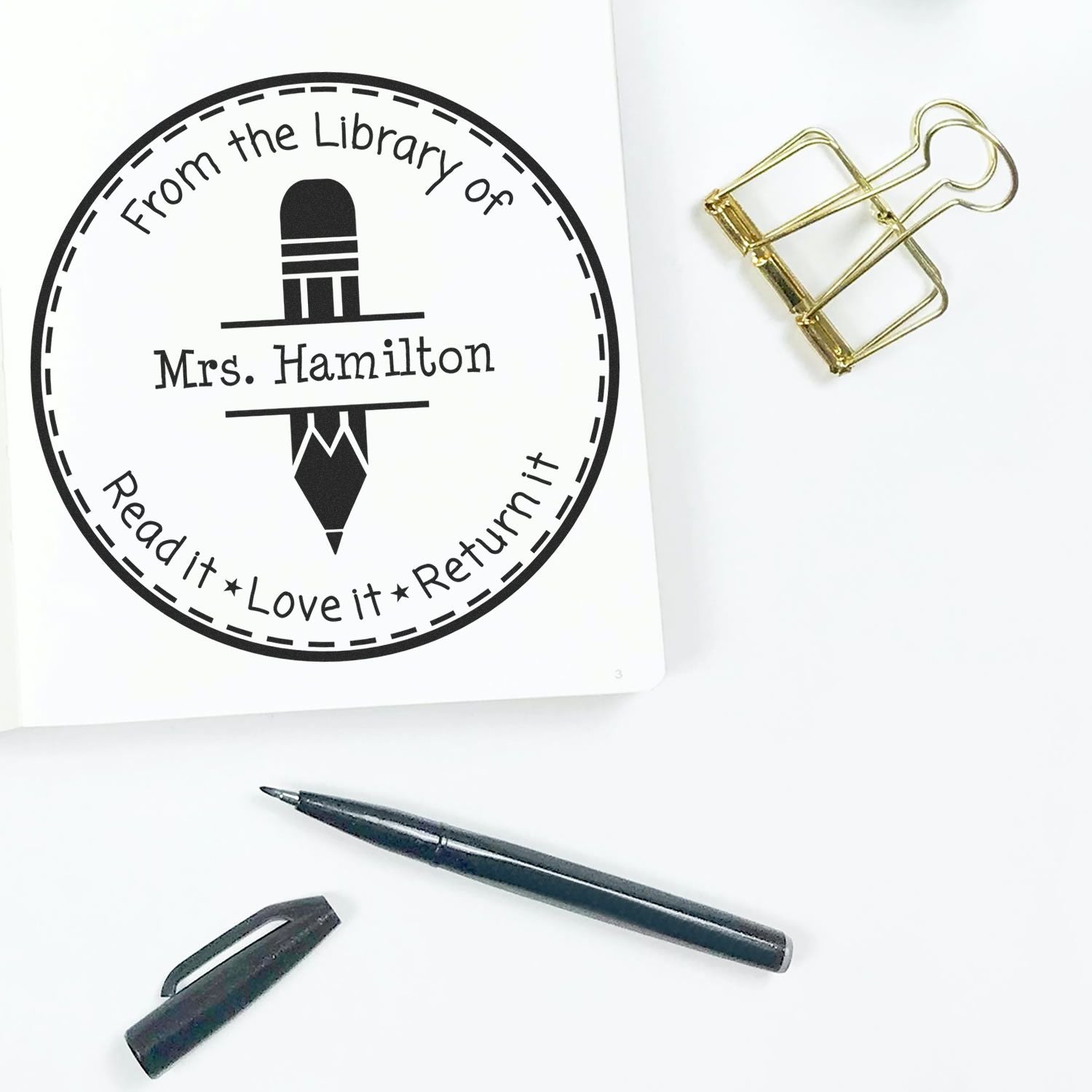 Round black library return stamp reading From the Library of Mrs. Hamilton, with pencil icon, gold binder clip, and black pen.