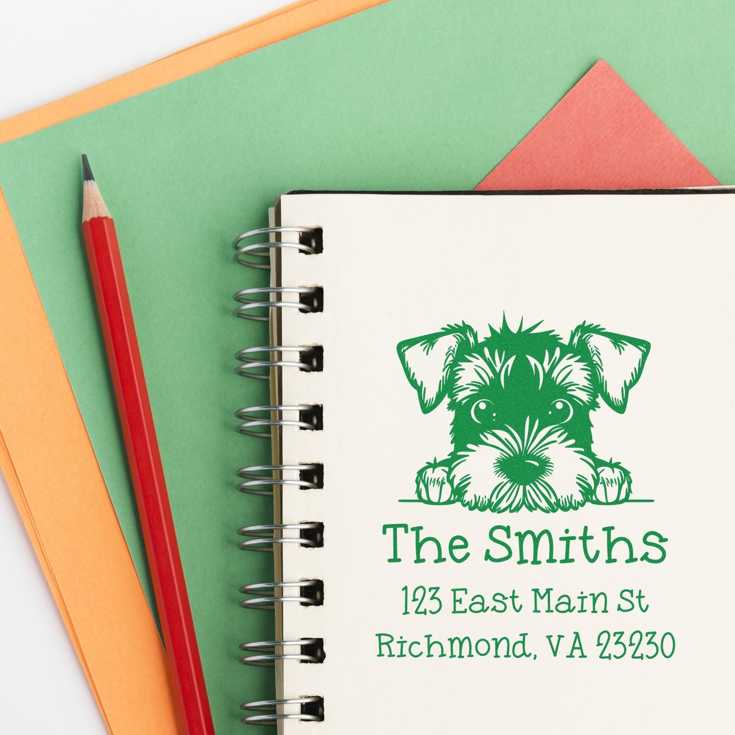 Green custom address stamp/seal with dog illustration on a spiral notebook, beside a red pencil on green, orange, and coral paper.