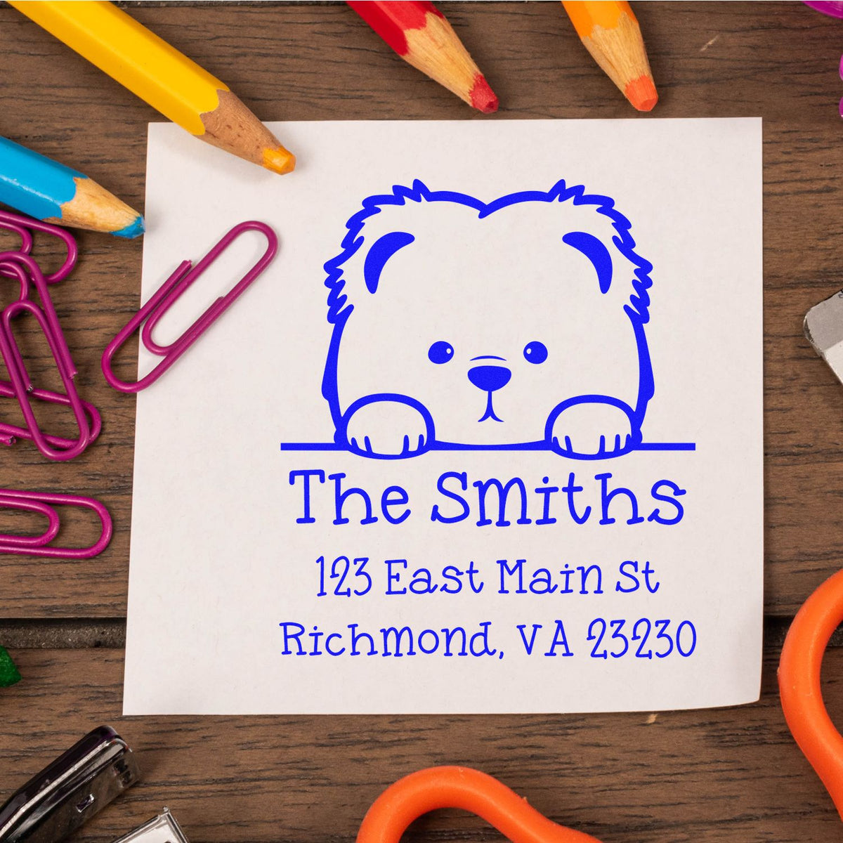 Blue custom stamp and seal with teddy bear logo and Smiths address on white paper, framed by colorful pencils and paper clips.