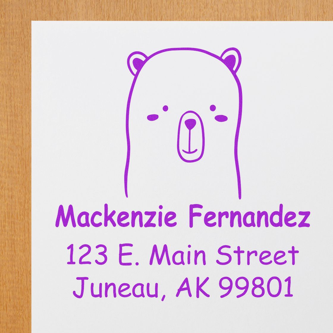 Wood handle custom stamp featuring an adorable bear cartoon animal design, made to order for mail use. The stamp is shown on a white paper with purple ink, displaying the name and address: Mackenzie Fernandez, 123 E. Main Street, Juneau, AK 99801.