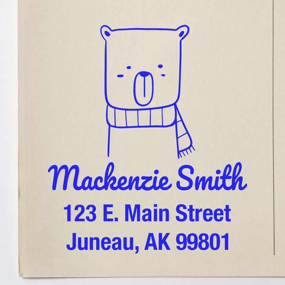 Wood handle snuggly bear cartoon animal made to order mail stamper in use. Personalized return mail stamp with a cute bear design, featuring the name "Mackenzie Smith" and address "123 E. Main Street, Juneau, AK 99801" in blue ink.