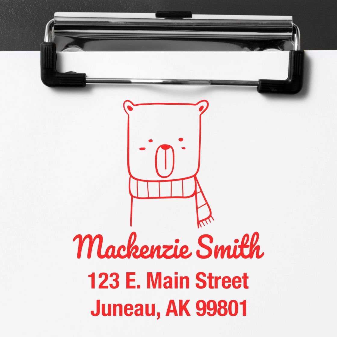 Wood handle snuggly bear cartoon animal made to order mail stamper on paper. Personalized home stamp with red bear design and address: Mackenzie Smith, 123 E. Main Street, Juneau, AK 99801, clipped to a clipboard.