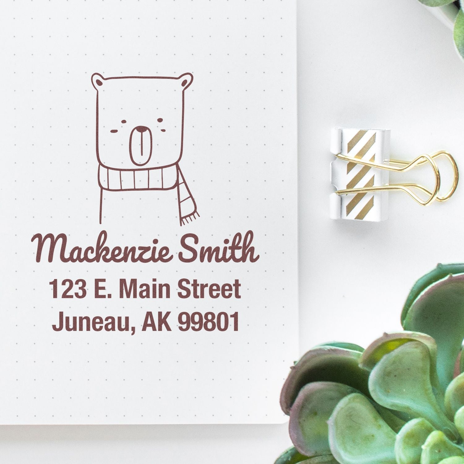 Custom address stamp featuring a snuggly bear cartoon with a wood handle. The mail stamper is made to order, displaying "Mackenzie Smith, 123 E. Main Street, Juneau, AK 99801" on dotted paper, next to a gold-striped binder clip and succulents.