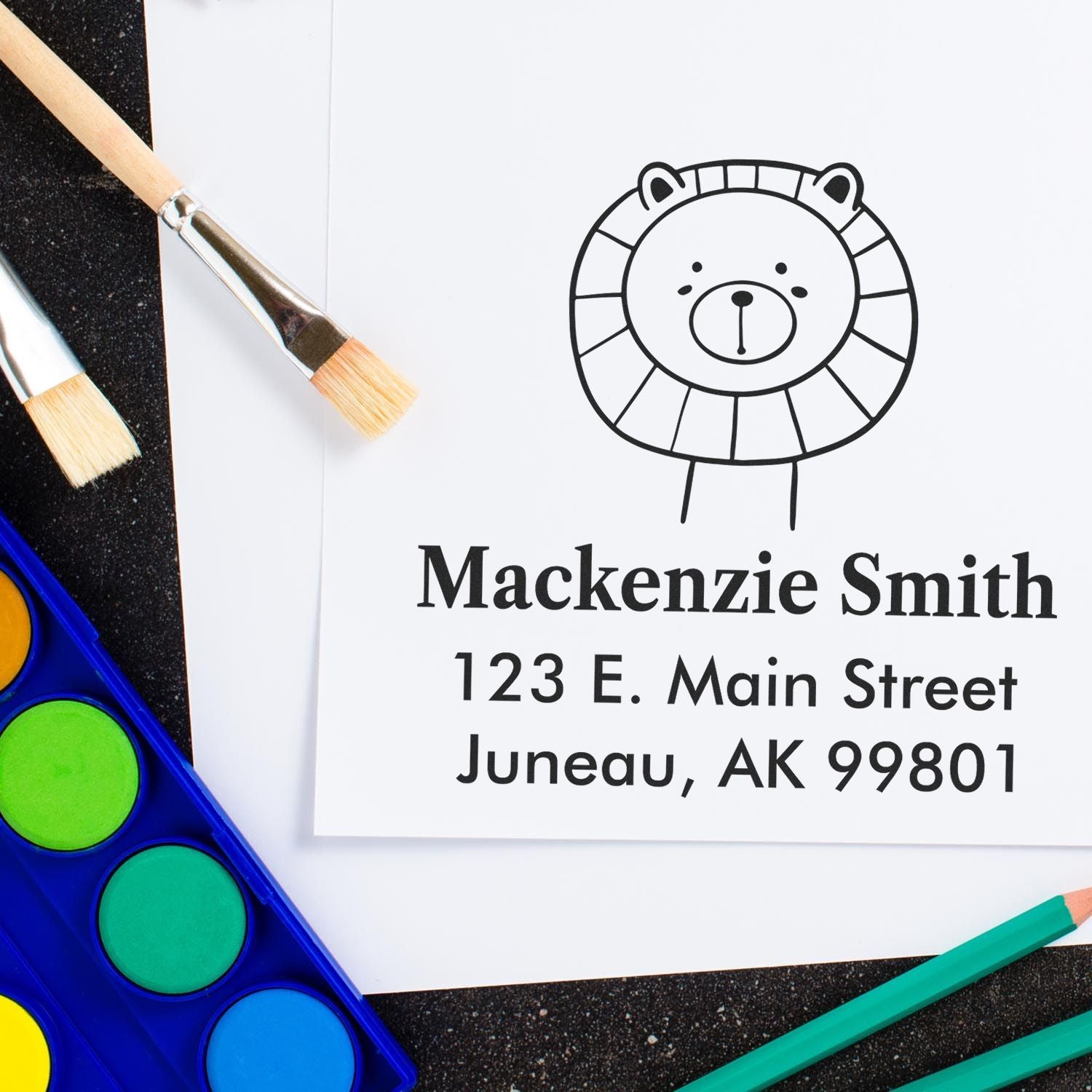 Self-Inking Cartoon Animal Lovable Lion's Lair Custom Made Mail Stamper on paper with "Mackenzie Smith, 123 E. Main Street, Juneau, AK 99801." Personalizable Address Stamper shown with paintbrushes and watercolor set nearby.