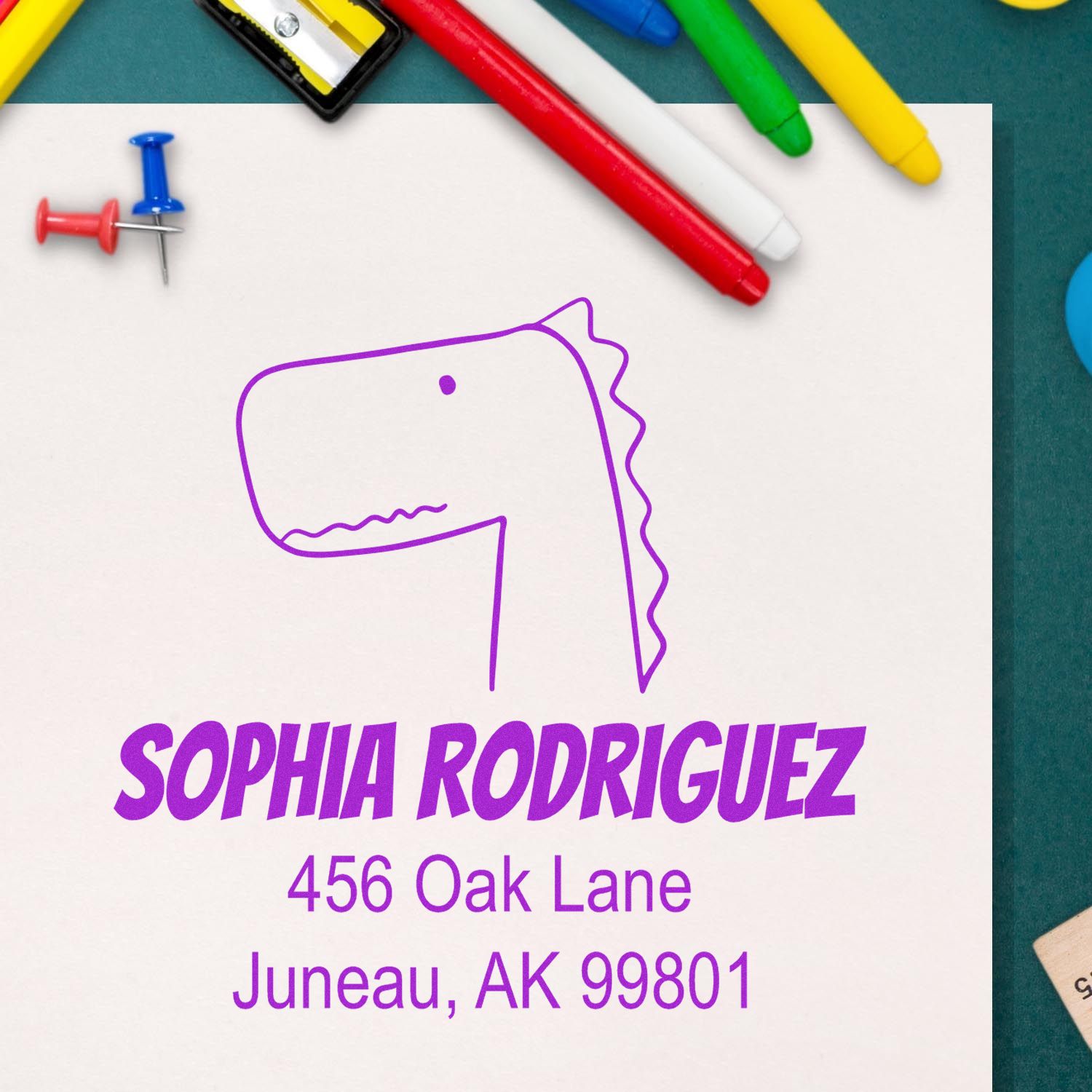 Self-Inking Cartoon Animal Mirthful Dino Custom Made Return Address Stamper on a white paper with the name "Sophia Rodriguez" and address "456 Oak Lane, Juneau, AK 99801" in purple. Surrounding items include colorful pens and push pins. Custom-Made Address Stamp.