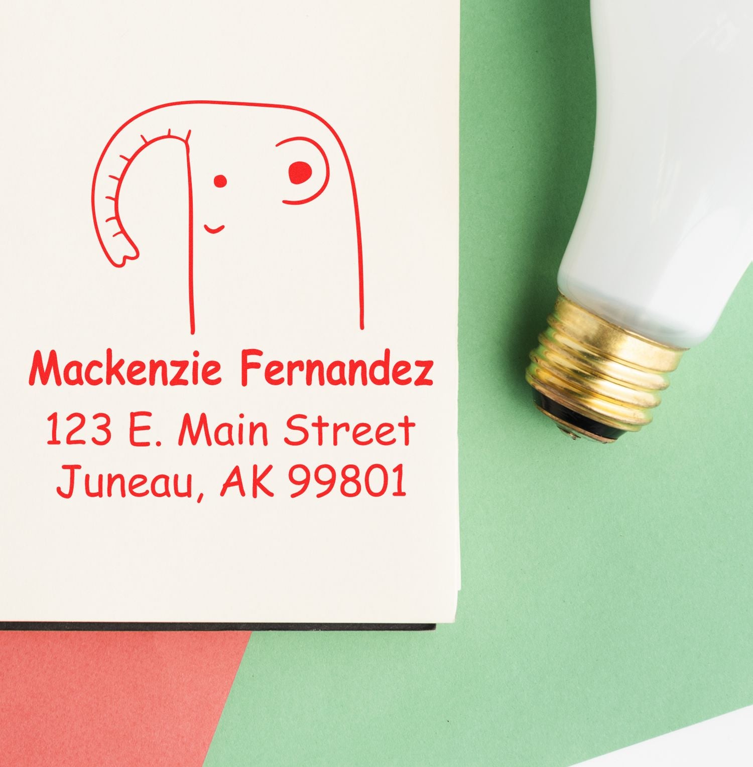 Wood handle endearing elephant cartoon animal made to order address stamper on a custom-made home stamp. The stamp features a cute red elephant design with personalized address details on a card, placed next to a light bulb on colorful paper.