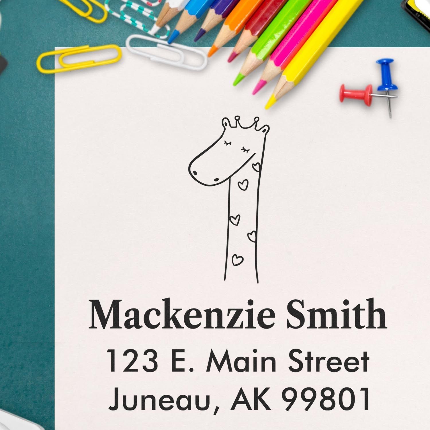 Portable Pre-Inked Lovely Giraffe Custom Designed Address Stamp on paper with "Mackenzie Smith, 123 E. Main Street, Juneau, AK 99801." Personalized Address Stamper features a giraffe illustration. Surrounding items include colored pencils, paper clips, and a push pin.