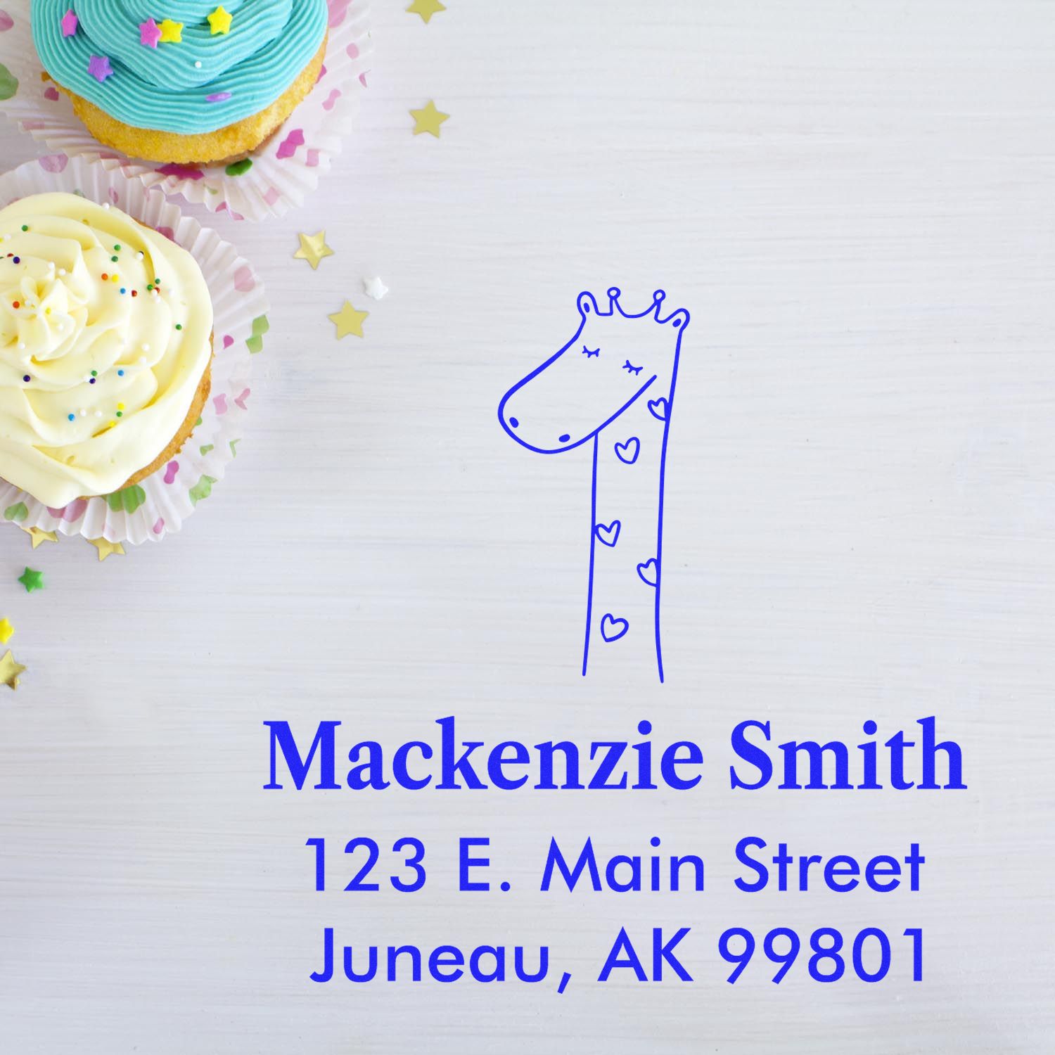 Self-inking cartoon animal lovely giraffe custom-made mailing stamp on a white surface. The custom-made address stamp features a cute giraffe design with hearts, alongside the name "Mackenzie Smith" and an address in Juneau, AK. Cupcakes with sprinkles are nearby.