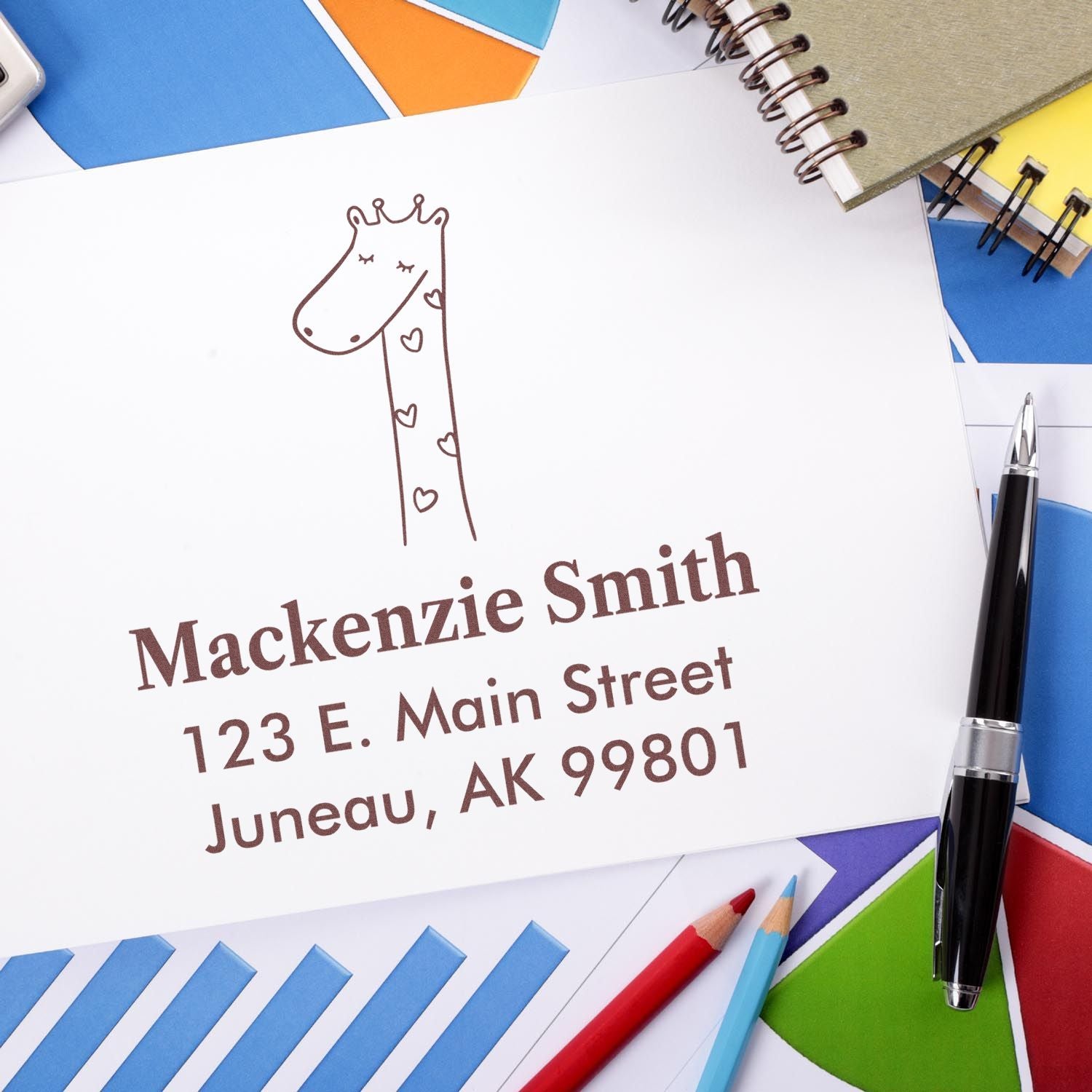 Self-inking cartoon animal lovely giraffe custom-made mailing stamp on an envelope with "Mackenzie Smith, 123 E. Main Street, Juneau, AK 99801." Surrounded by colorful papers, a pen, and a notebook, showcasing a custom-made home stamp design.