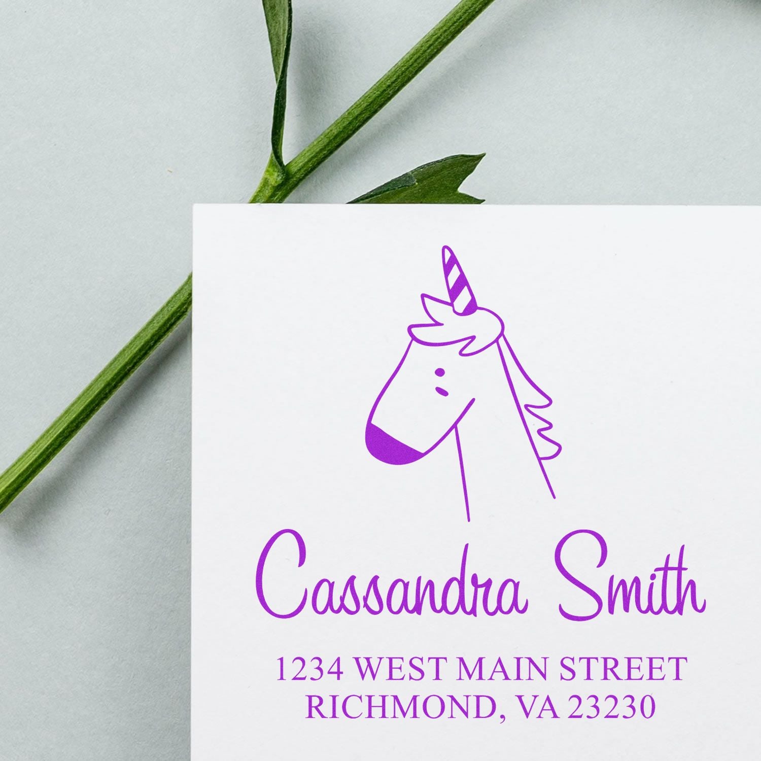 Wood handle whimsical unicorn cartoon animal made to order address stamp on an envelope. Customizable return mail stamp with purple ink, featuring a unicorn design and personalized address details. A green stem lies beside the envelope.