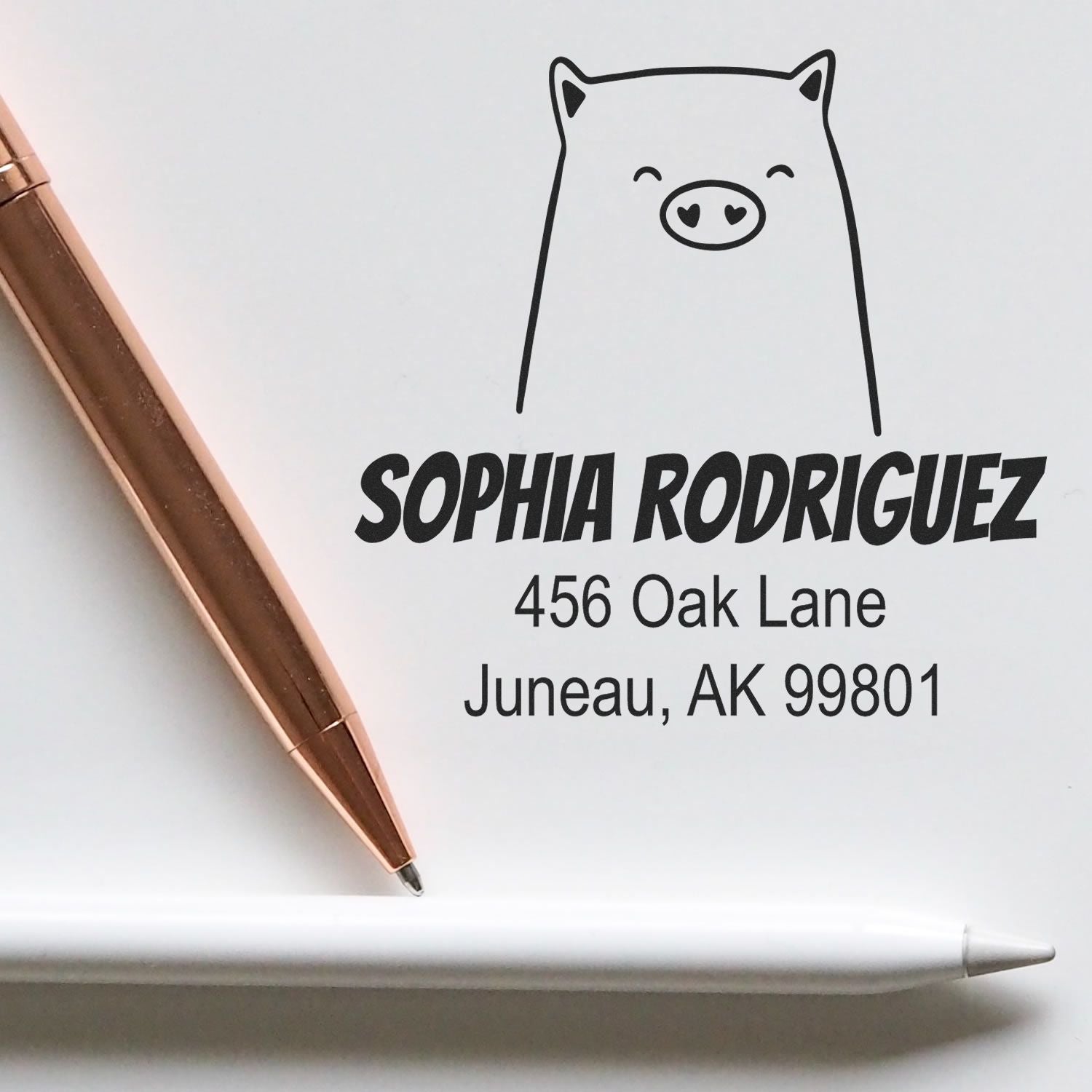 Self-inking cartoon animal cheery pig custom made address stamp on white paper with the name "Sophia Rodriguez" and address "456 Oak Lane, Juneau, AK 99801" in bold. A gold pen and a white pen are placed beside the custom address stamp.