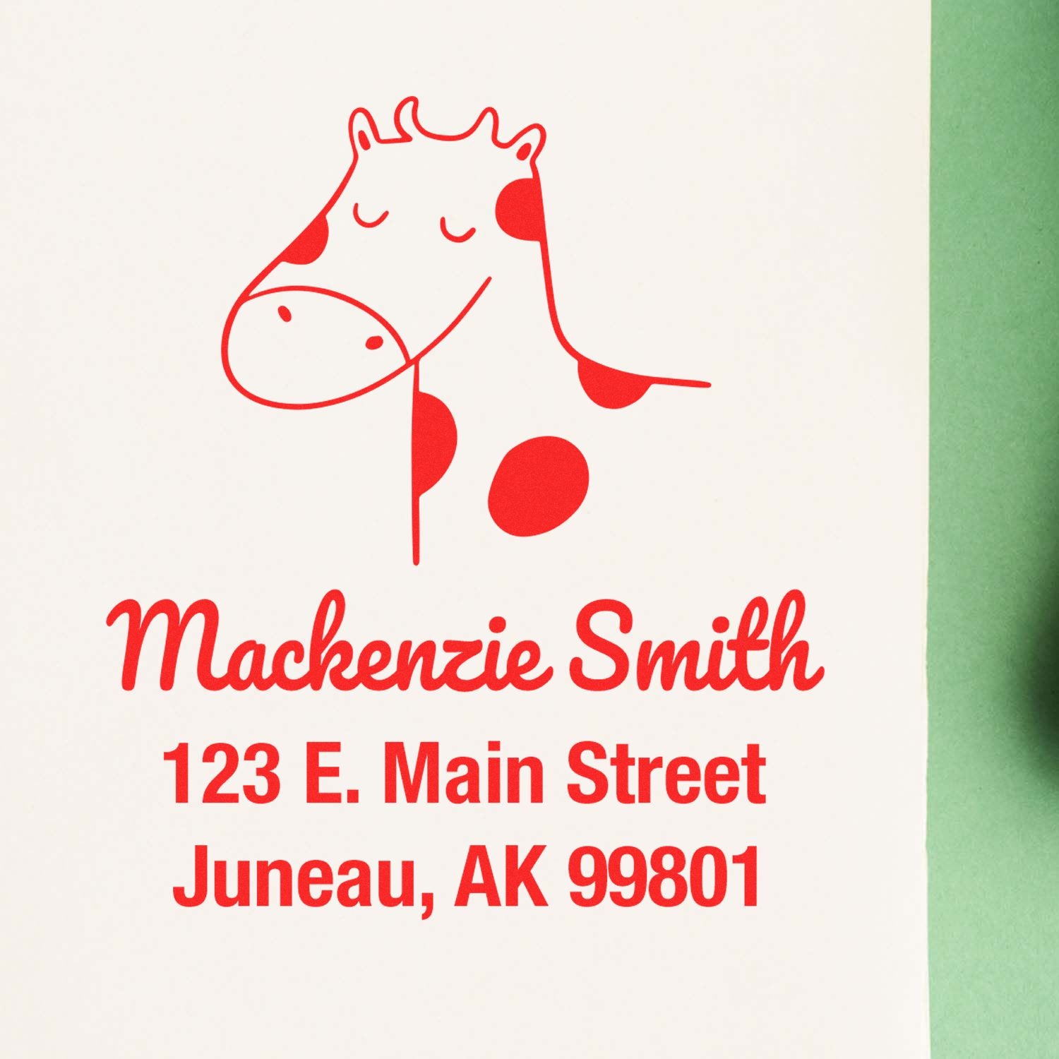 Image of a Cow Cuddles Pre-Inked Customized Address Stamper imprint on paper, featuring a cute cow illustration above the text "Mackenzie Smith, 123 E. Main Street, Juneau, AK 99801." This Personalizable Home Stamp is shown on a green background.