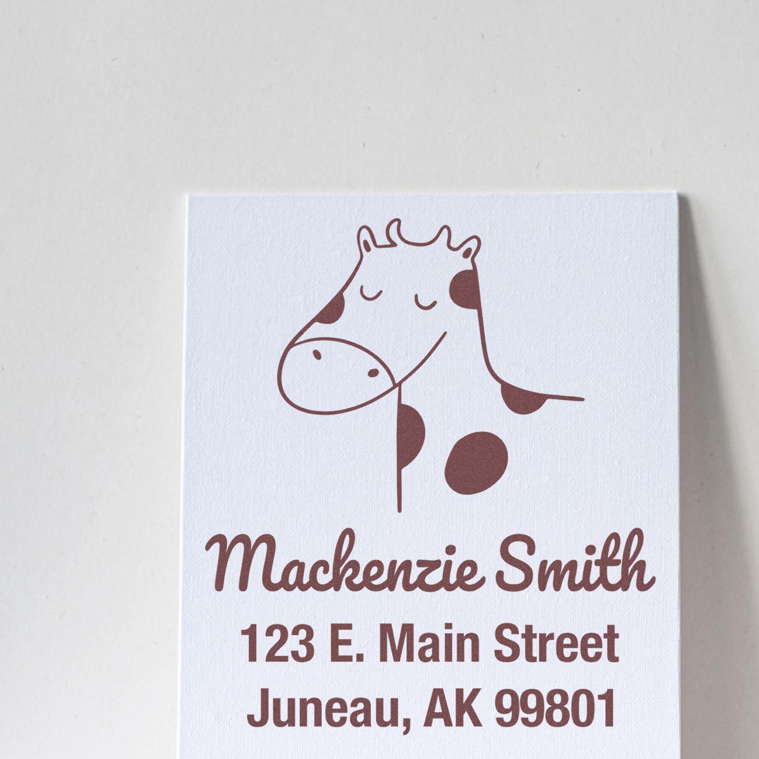 Self-Inking Cartoon Animal Cow Cuddles Custom Made Home Stamp featuring a smiling cow design. Personalized Return Mail Stamp with the name "Mackenzie Smith" and address "123 E. Main Street, Juneau, AK 99801" in brown ink on white paper.
