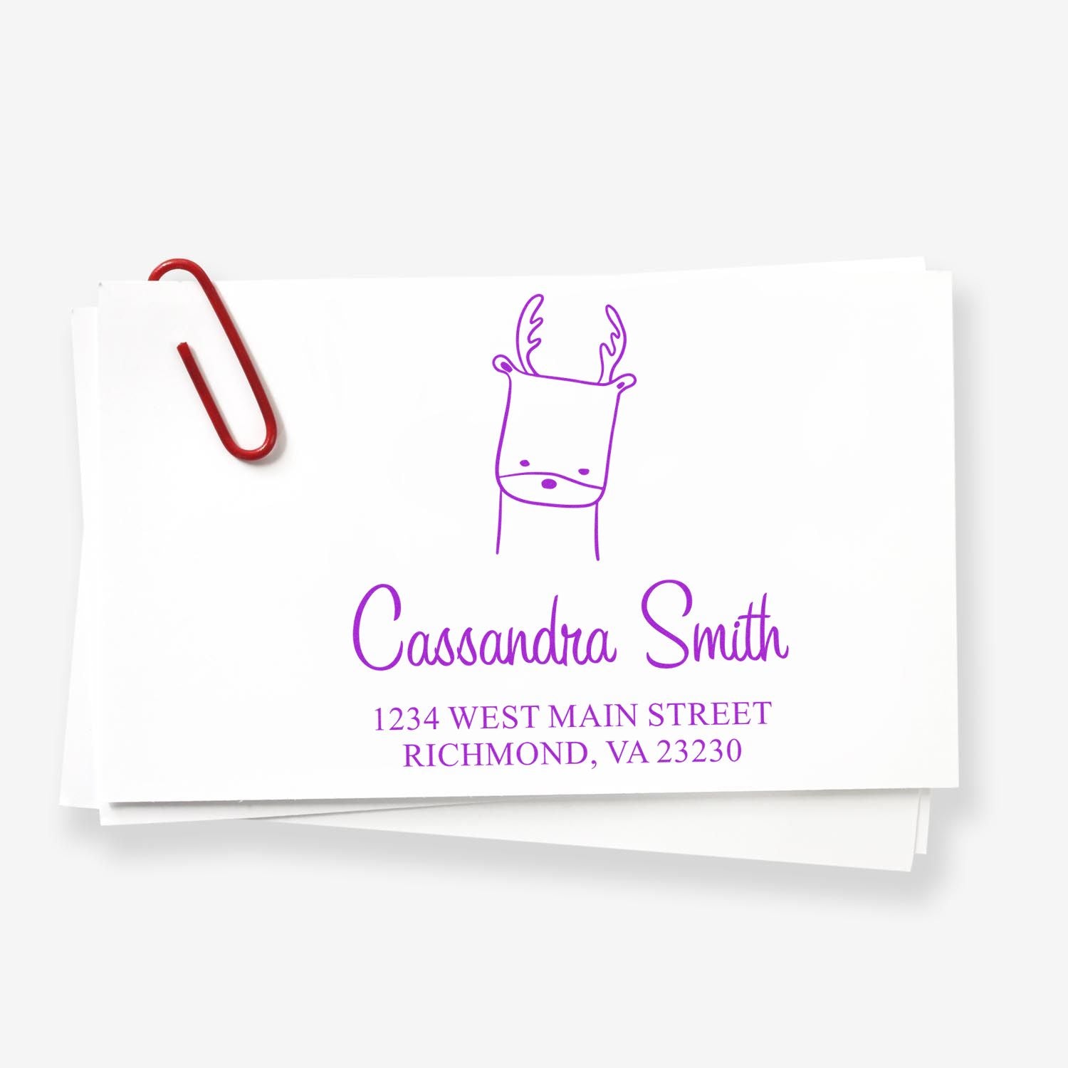 Wood handle regal reindeer cartoon animal made to order envelope stamper displayed on a stack of envelopes. The personalized address stamp features a purple reindeer design with the name "Cassandra Smith" and an address in Richmond, VA.