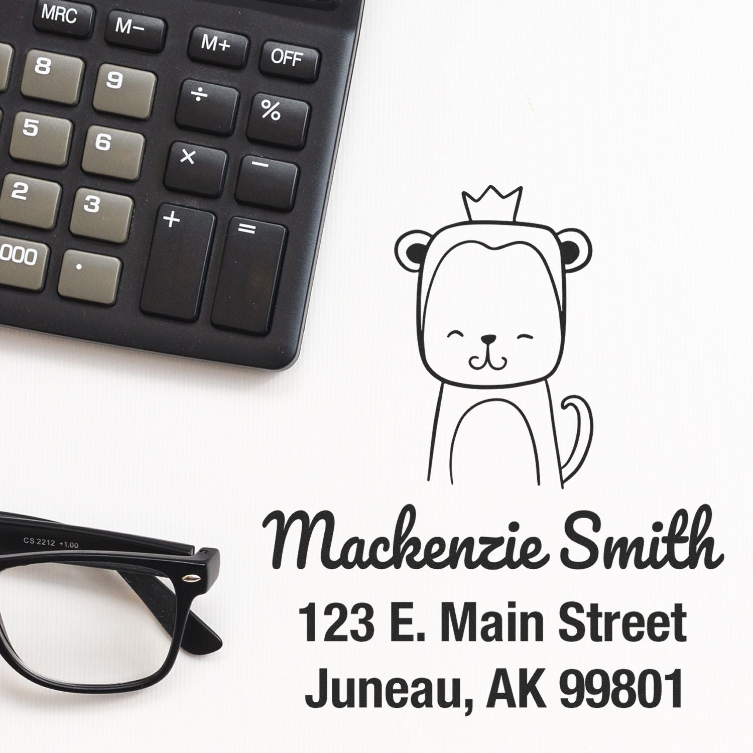 Wood handle mischievous monkey cartoon animal made to order name and address stamp displayed next to a calculator and glasses. Customizable address stamp with "Mackenzie Smith, 123 E. Main Street, Juneau, AK 99801" printed on paper.
