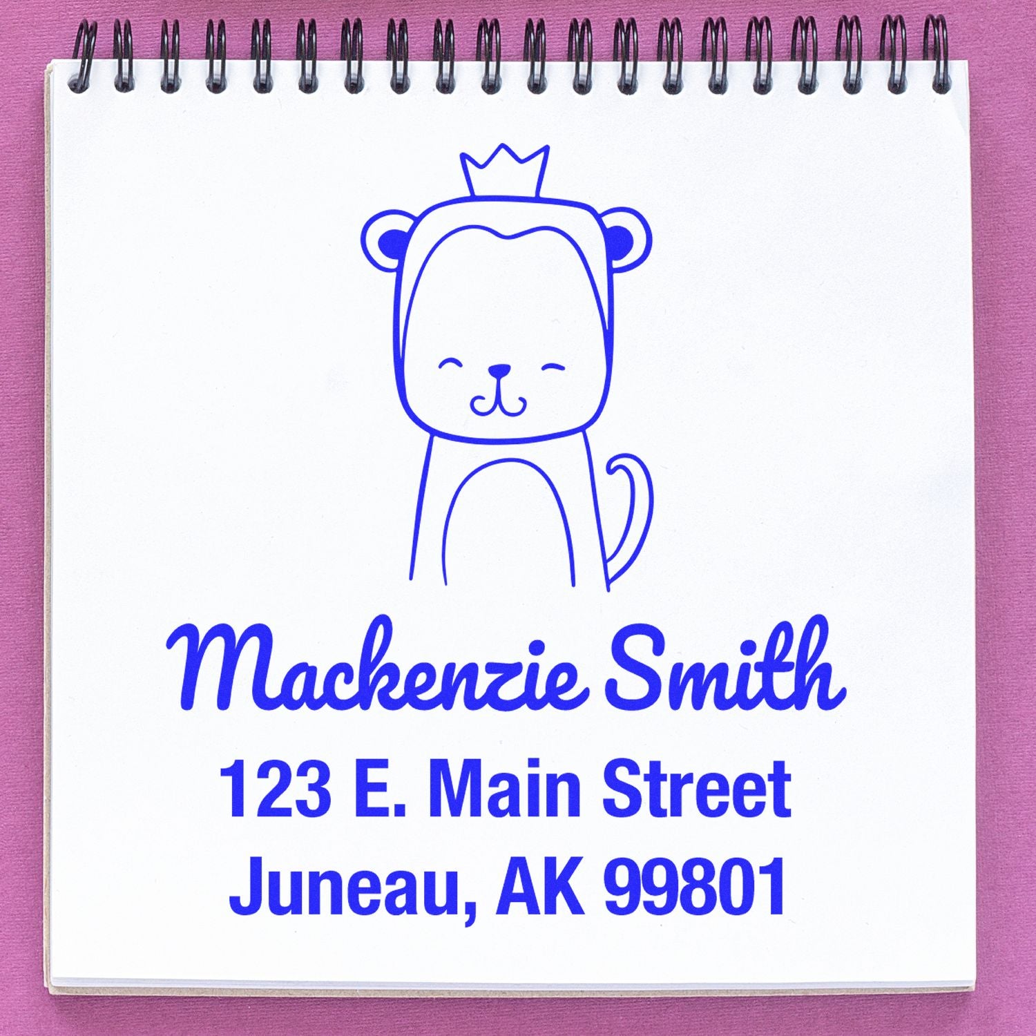 Mischievous Monkey Pre-Inked Customized Address Stamp featuring a cute monkey with a crown, used as a Personalized Return Mail Stamper. The stamp displays the name "Mackenzie Smith" and address "123 E. Main Street, Juneau, AK 99801" on a spiral notebook.