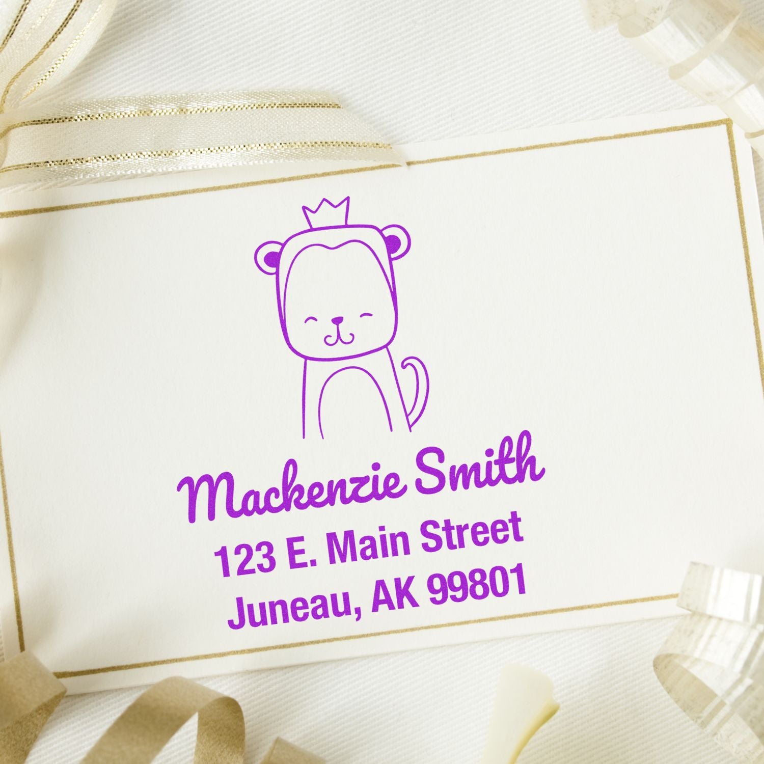 Wood handle mischievous monkey cartoon animal made to order name and address stamp on a card. Customized return mail stamp with purple text showing "Mackenzie Smith, 123 E. Main Street, Juneau, AK 99801" surrounded by ribbon.