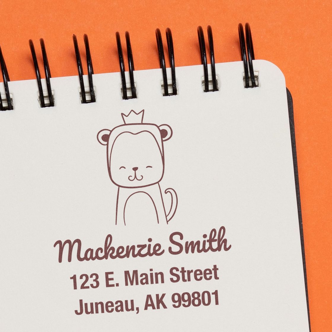 Portable Pre-Inked Mischievous Monkey Custom Designed Envelope Stamper in use on a notebook. The Custom Return Mail Stamper features a playful monkey illustration with a crown, and personalized text for Mackenzie Smith, 123 E. Main Street, Juneau, AK 99801.