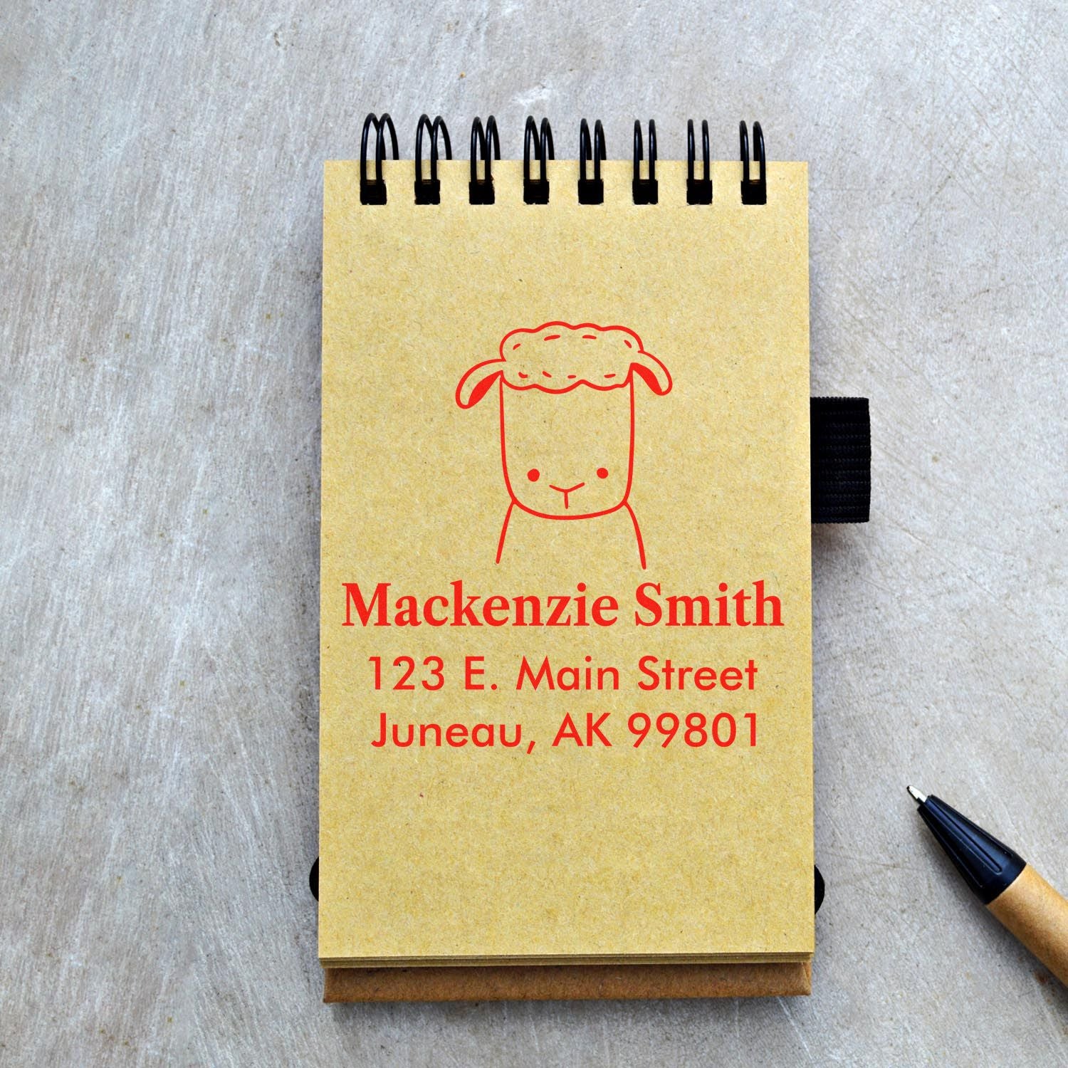 A notebook with a red Fluffy Sheep Pre-Inked Customized Address Stamper imprint showing "Mackenzie Smith, 123 E. Main Street, Juneau, AK 99801." A pen lies nearby. This Custom-Made Return Mail Stamper adds a personal touch to correspondence.