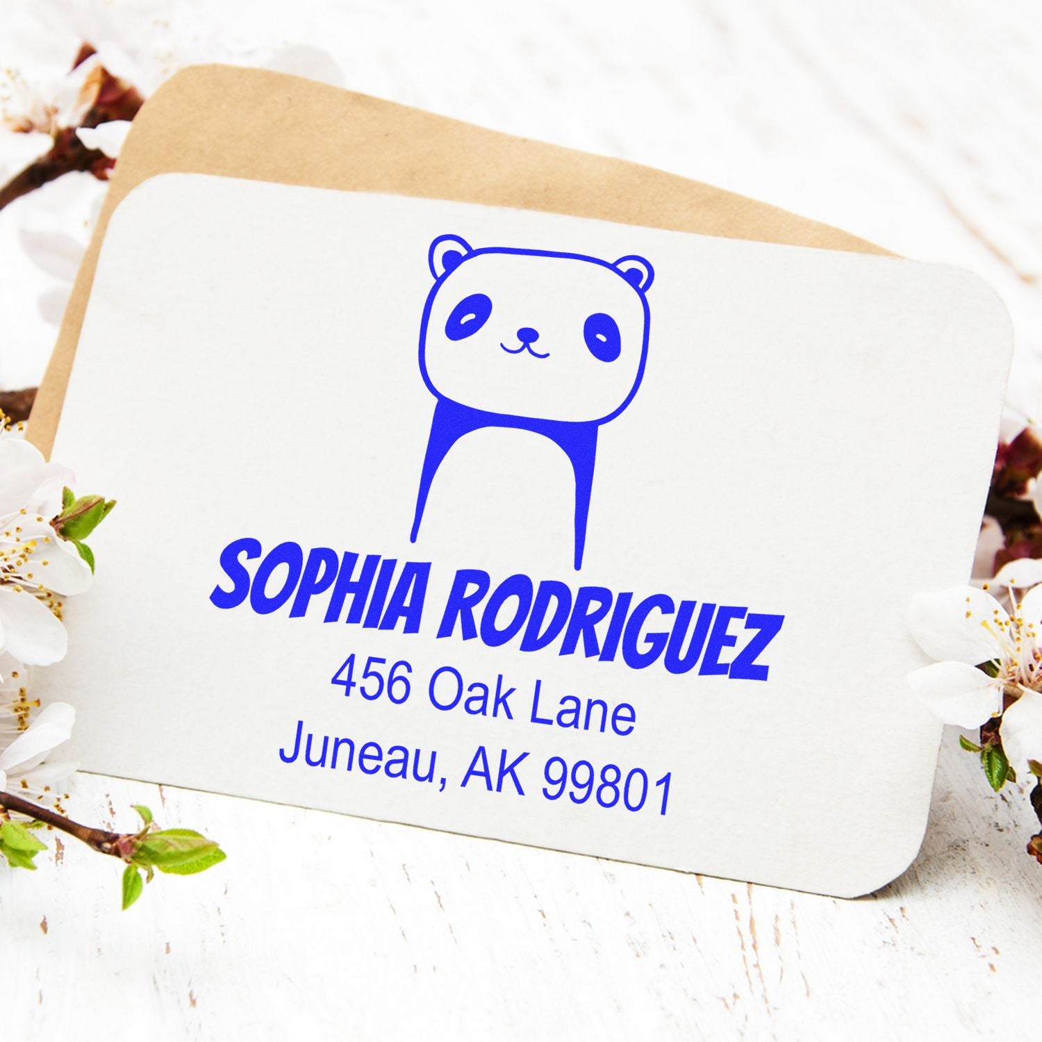 Playful Panda Pre-Inked Customized Name And Address Stamper on a card with "Sophia Rodriguez, 456 Oak Lane, Juneau, AK 99801" in blue text. The Custom New Address Stamp features a cute panda design, surrounded by white flowers on a wooden surface.