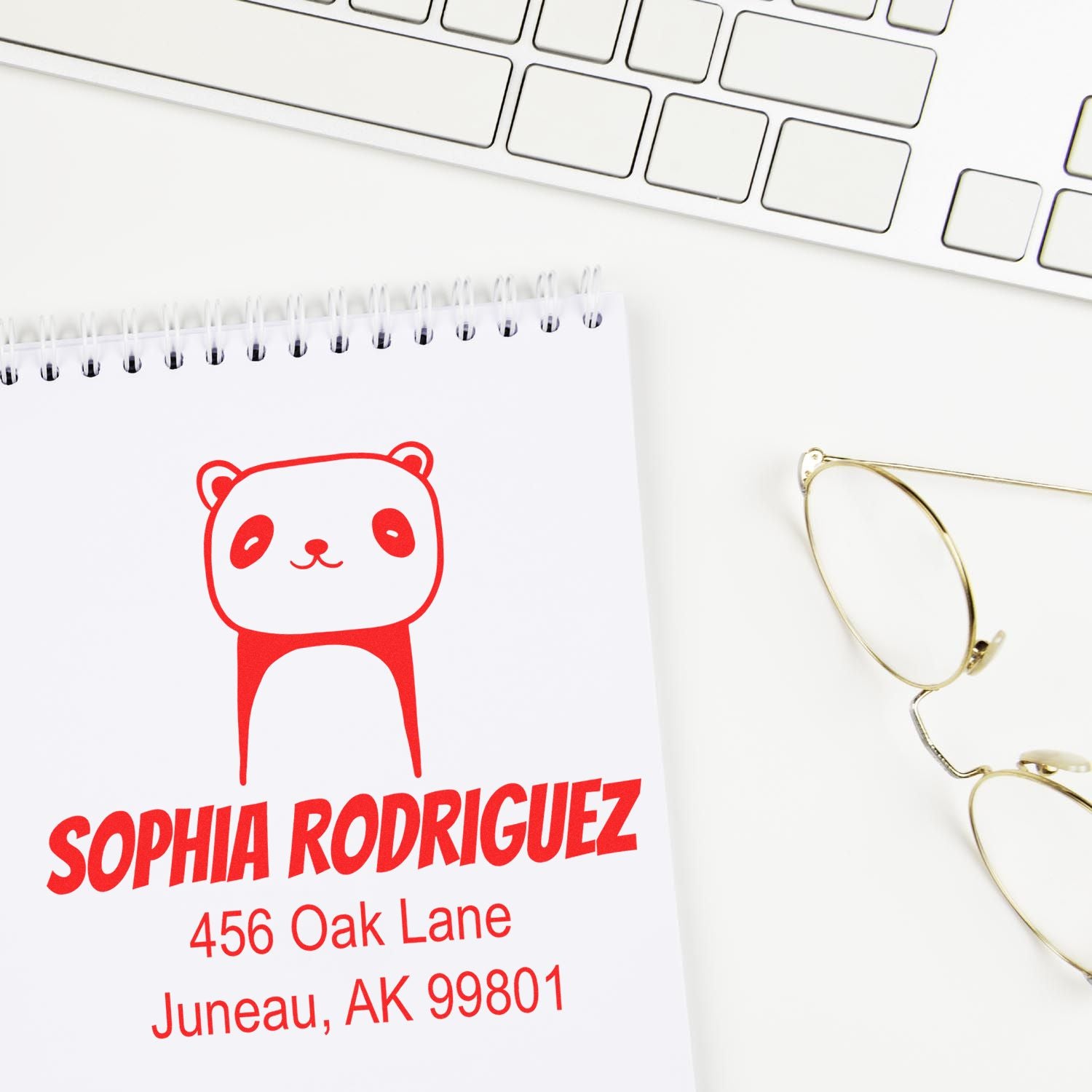 Portable Pre-Inked Playful Panda Custom Designed Name And Address Stamp on a notepad with Sophia Rodriguez, 456 Oak Lane, Juneau, AK 99801 in red ink. Customizable Mailing Stamper next to a keyboard and glasses.
