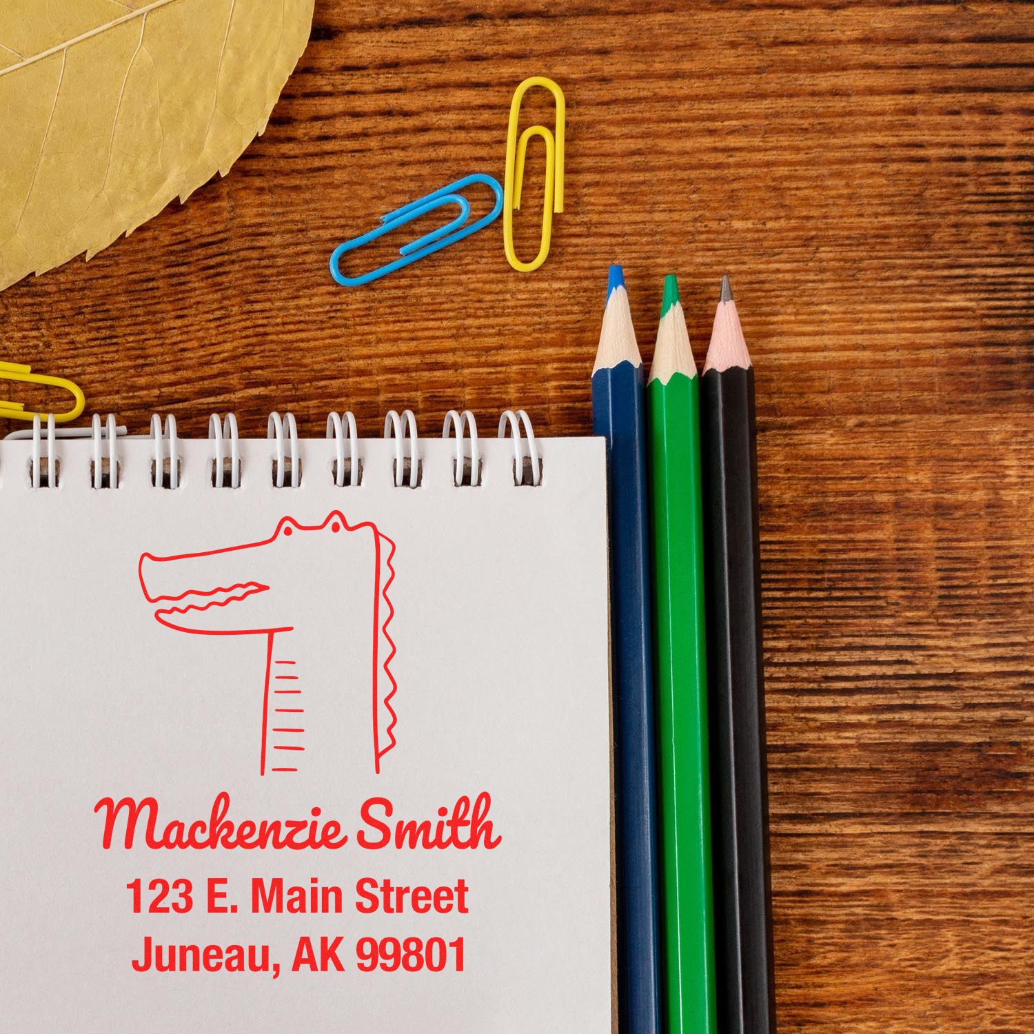 Wood handle bright-eyed crocodile cartoon animal made to order home address stamper on a notepad. Customized home stamp with red ink shows name and address. Nearby are colored pencils, paper clips, and a leaf on a wooden surface.