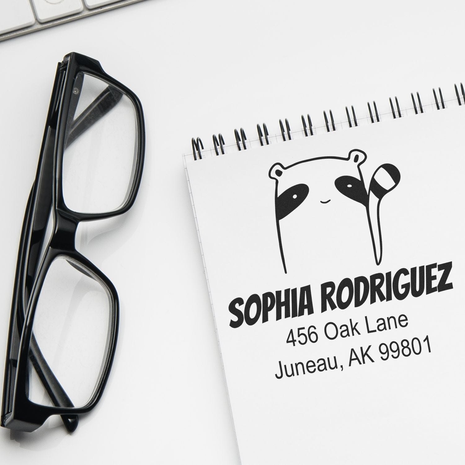 A Renegade Racoon Pre-Inked Customized Return Address Stamp is shown on a notepad with the name "Sophia Rodriguez" and address "456 Oak Lane, Juneau, AK 99801." Glasses and a keyboard are nearby. This Custom Return Mail Stamp features a raccoon design.