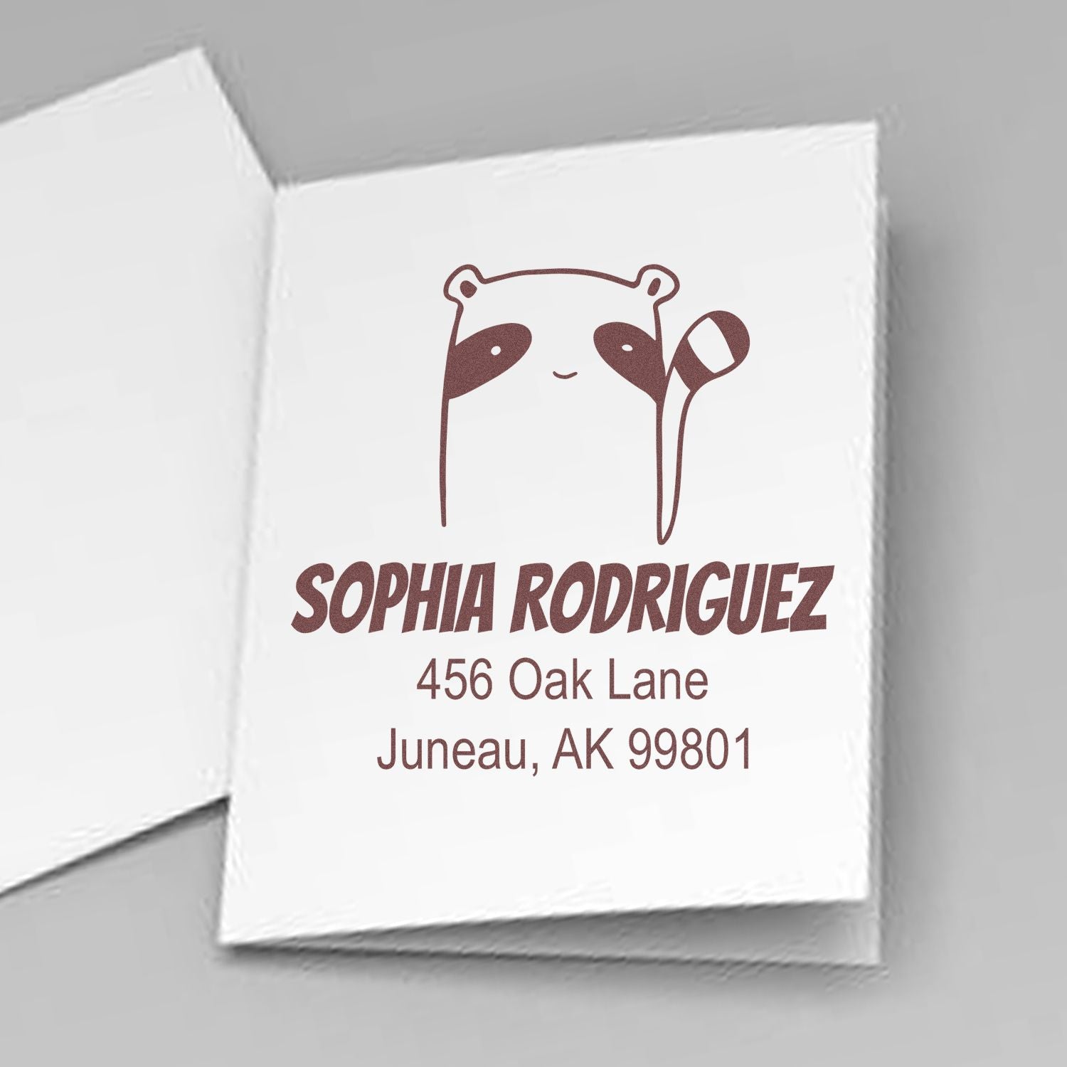 Self-Inking Cartoon Animal Renegade Racoon Custom Made Return Address Stamper displayed on a card. The design features a raccoon with text "Sophia Rodriguez, 456 Oak Lane, Juneau, AK 99801." Personalizable New Address Stamp for unique mailing needs.