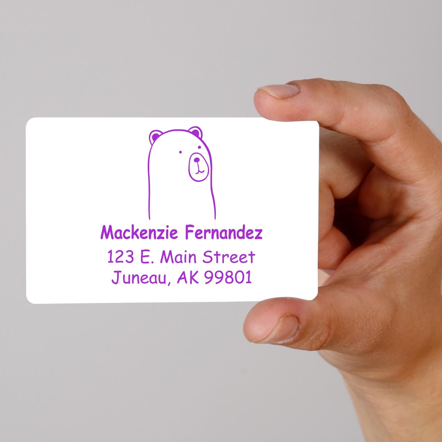 A hand holds a card stamped with a Portable Pre-Inked Cuddly Polar Bear Custom Designed Return Address Stamp. The purple bear design is above the text "Mackenzie Fernandez, 123 E. Main Street, Juneau, AK 99801." This Personalizable New Address Stamper adds charm to mail.