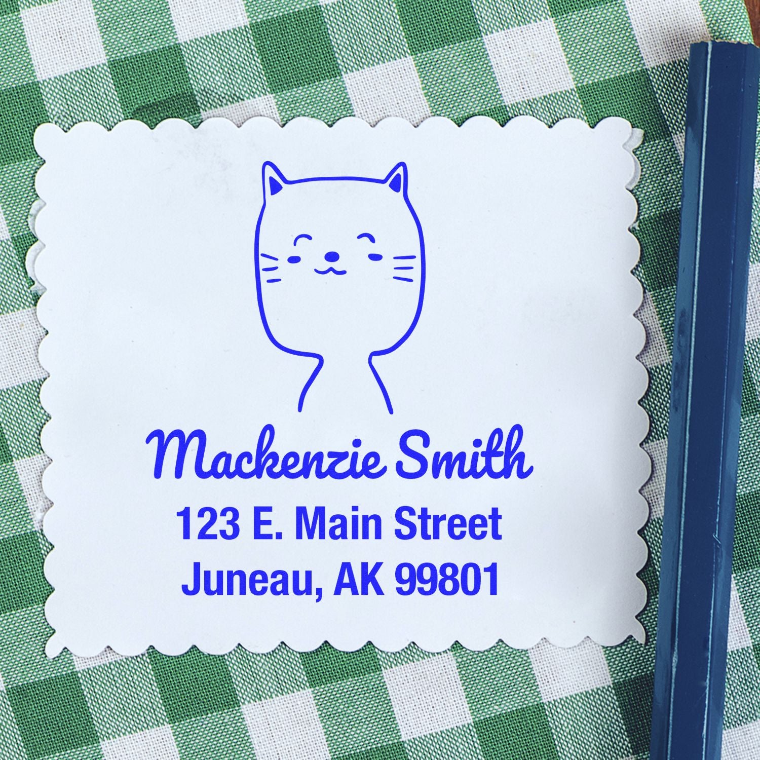 Charming Cat Pre-Inked Customized New Home Stamp on a white card with scalloped edges, featuring a cute cat illustration and personalized return mail stamp details: "Mackenzie Smith, 123 E. Main Street, Juneau, AK 99801," placed on a green checkered cloth.
