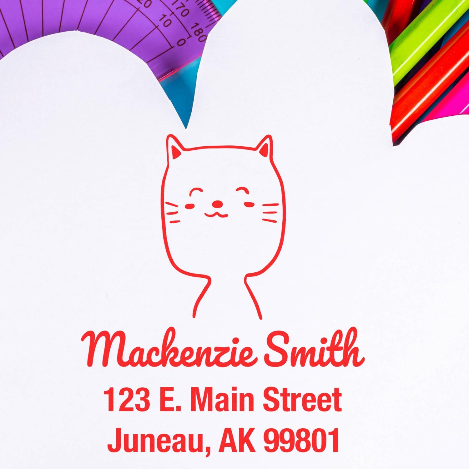 Wood handle custom mail stamp featuring a charming cat cartoon animal design. The stamp is used on an envelope with the text "Mackenzie Smith, 123 E. Main Street, Juneau, AK 99801." Includes colorful stationery in the background.
