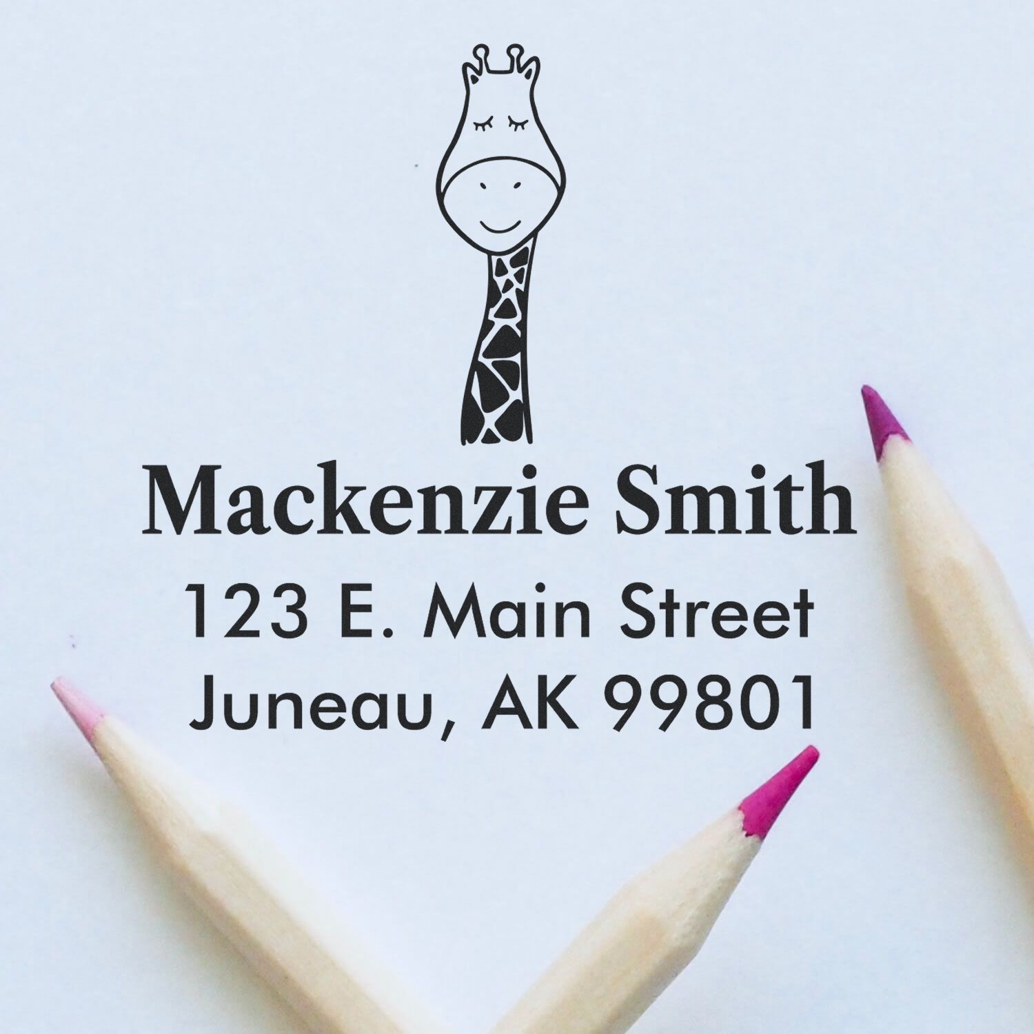 Wood Handle Jolly Giraffe Cartoon Animal Custom Mail Stamper imprint on paper, featuring a smiling giraffe above the name "Mackenzie Smith" and address. Two pencils with pink tips are placed nearby. Custom Mailing Stamp design.