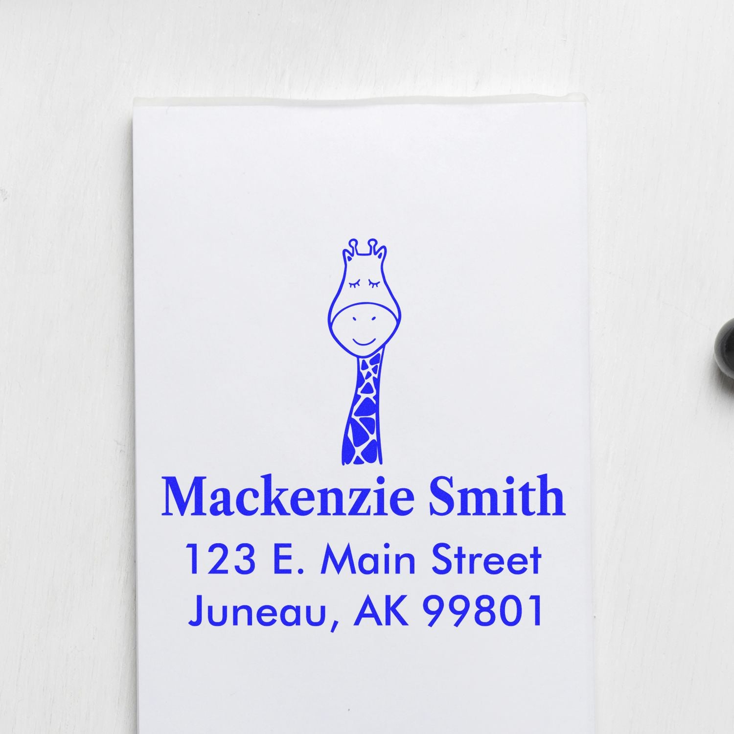 Portable Pre-Inked Jolly Giraffe Custom Designed New Home Stamp on white paper, featuring a smiling giraffe illustration above personalized mailing details: "Mackenzie Smith, 123 E. Main Street, Juneau, AK 99801." Personalized Mailing Stamp in blue ink.