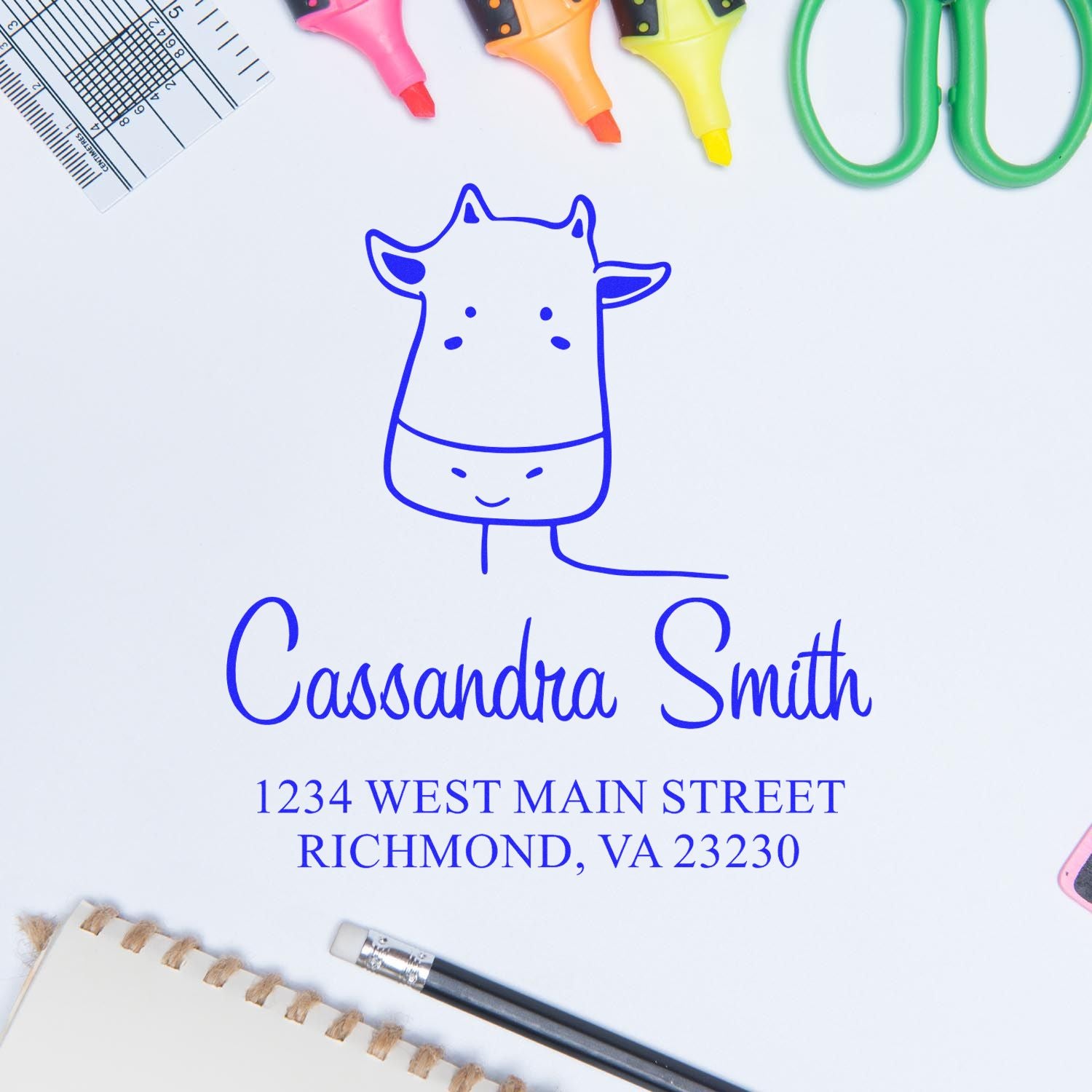 Portable Pre-Inked Giraffe Grins Custom Designed New Home Stamper on a desk with stationery. The Personalizable New Address Stamp displays "Cassandra Smith, 1234 West Main Street, Richmond, VA 23230" in blue ink, surrounded by colorful office supplies.
