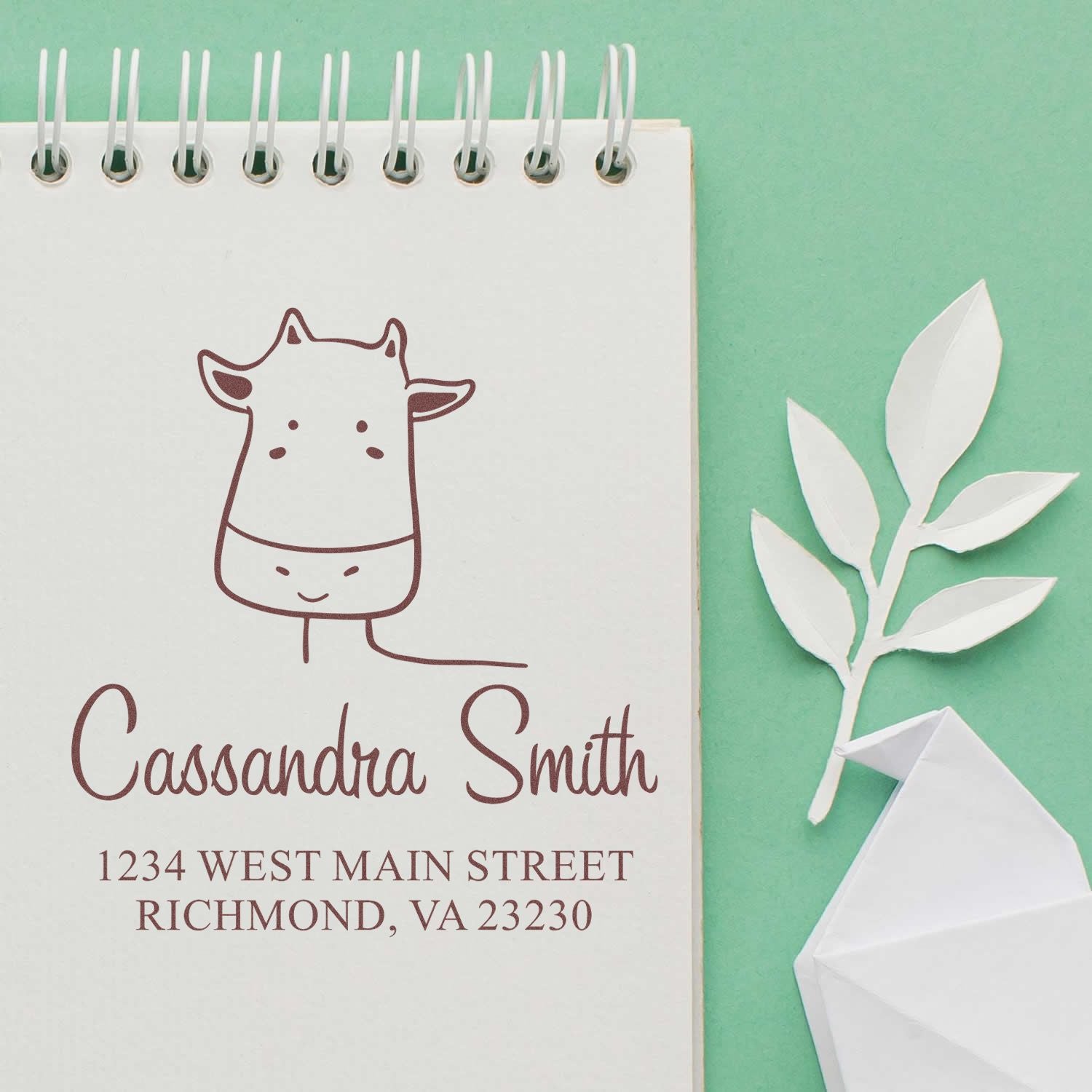 A Portable Pre-Inked Giraffe Grins Custom Designed New Home Stamper is shown on a notepad. The Customizable Mailing Stamp features a cute giraffe illustration with the name Cassandra Smith and an address in Richmond, VA. A paper leaf decorates the scene.
