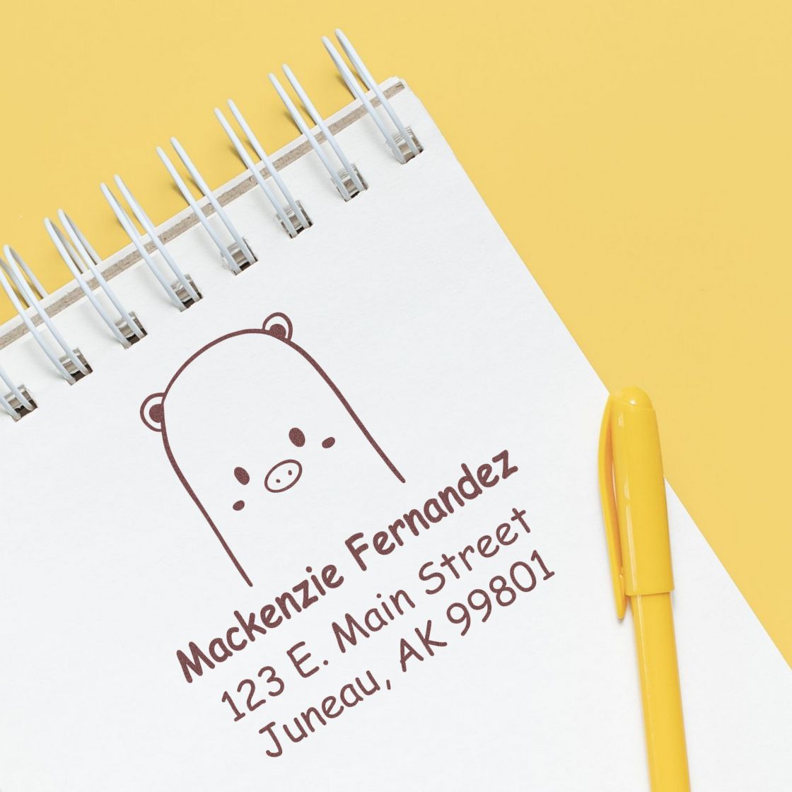 Self-inking cartoon animal beautiful bear personalized return address stamper on a notepad. The customized mailing stamp features a cute bear design with the name Mackenzie Fernandez and an address in Juneau, AK. A yellow pen lies beside the notepad.