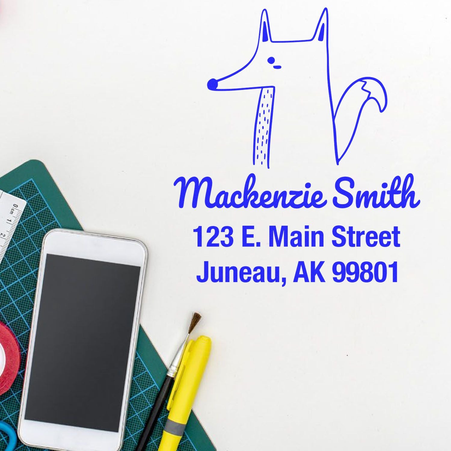 Wood Handle Finicky Fox Cartoon Animal Custom Address Stamper displayed on a desk with a phone, cutting mat, and stationery. The Customized Return Mail Stamp features a fox illustration and personalized address for Mackenzie Smith, 123 E. Main Street, Juneau, AK 99801.