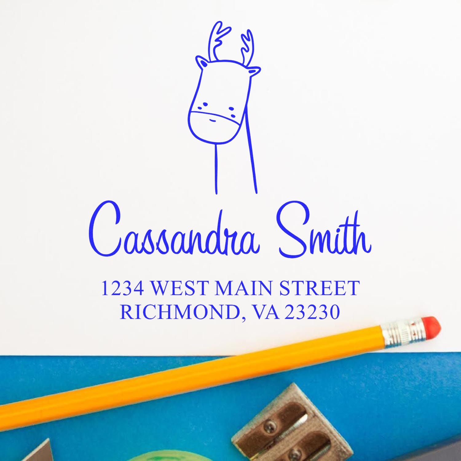 A Darling Deer Pre-Inked Handcrafted Address Stamper is shown in use, featuring a custom-made new address stamp with a cute deer illustration and the name "Cassandra Smith" alongside an address. A pencil and sharpener are nearby.