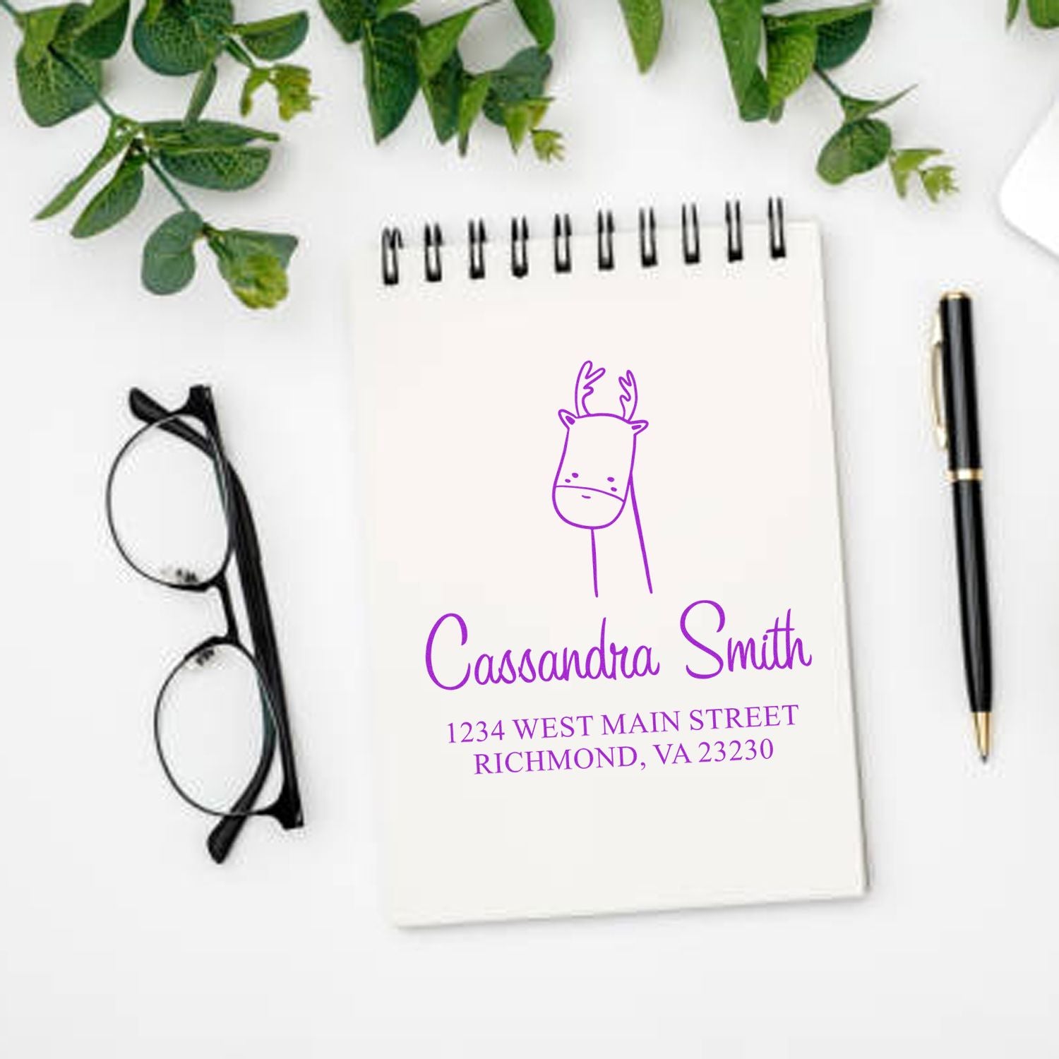 A notebook with a Darling Deer Pre-Inked Handcrafted Address Stamper design featuring a deer illustration and custom-made home stamp details for "Cassandra Smith, 1234 West Main Street, Richmond, VA 23230" in purple ink. Nearby are glasses and a pen.