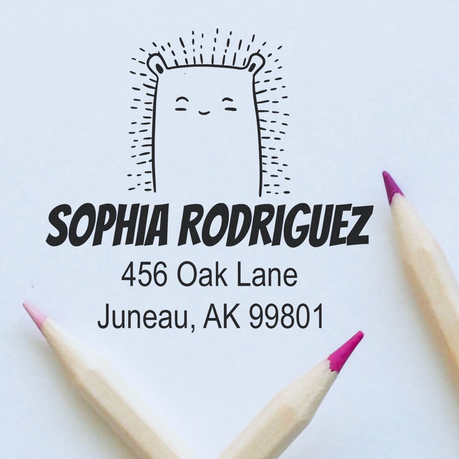 Image of a Portable Pre-Inked Bouncy Bear Made To Order Address Stamper on paper, featuring a cute bear design with the name "Sophia Rodriguez" and address "456 Oak Lane, Juneau, AK 99801." Two colored pencils are placed nearby. Custom Address Stamper in use.