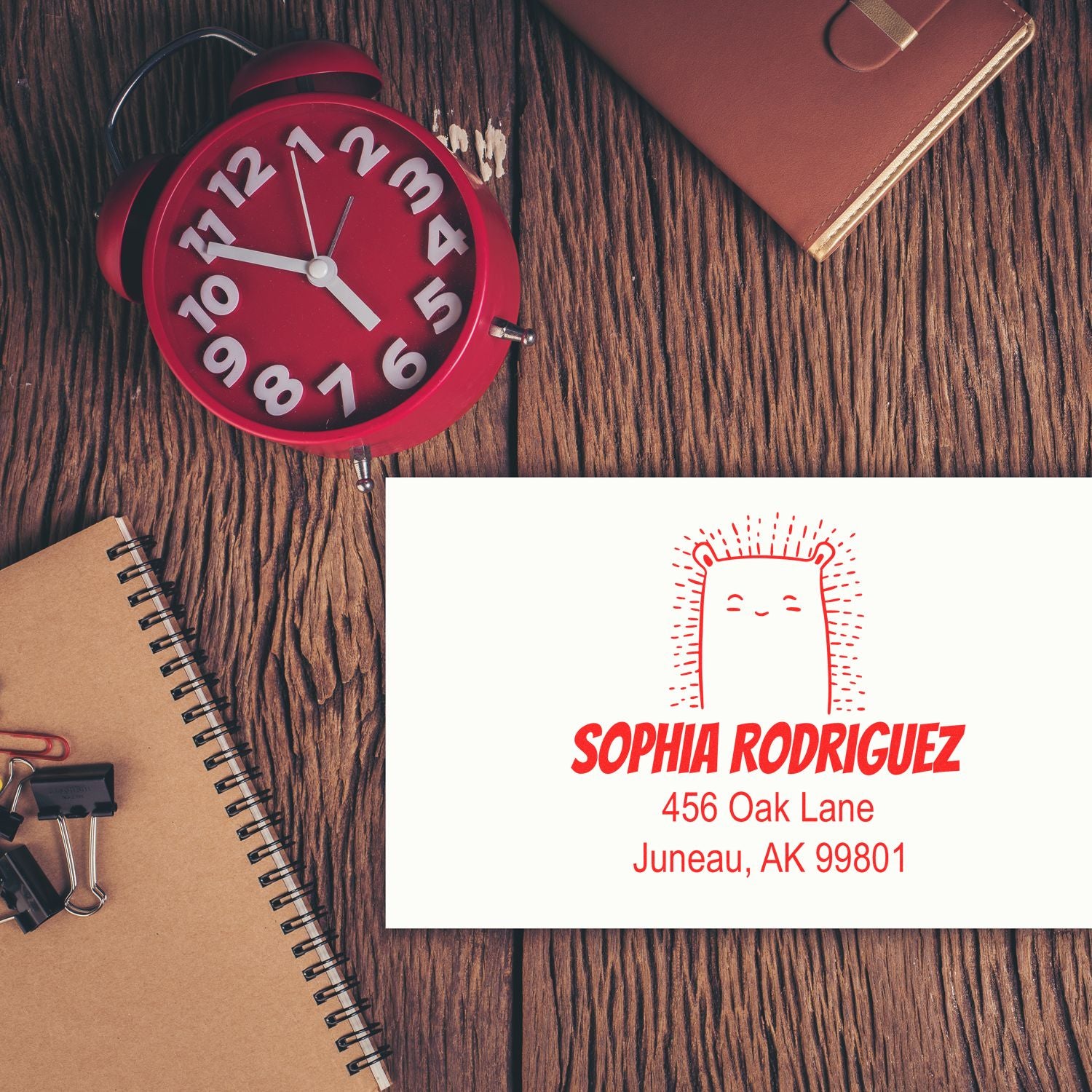 Wood Handle Bouncy Bear Cartoon Animal Custom Address Stamp on a white envelope with red ink, featuring customizable mailing stamp details for Sophia Rodriguez, 456 Oak Lane, Juneau, AK 99801. Nearby are a red clock, notebook, and paper clips on a wooden surface.