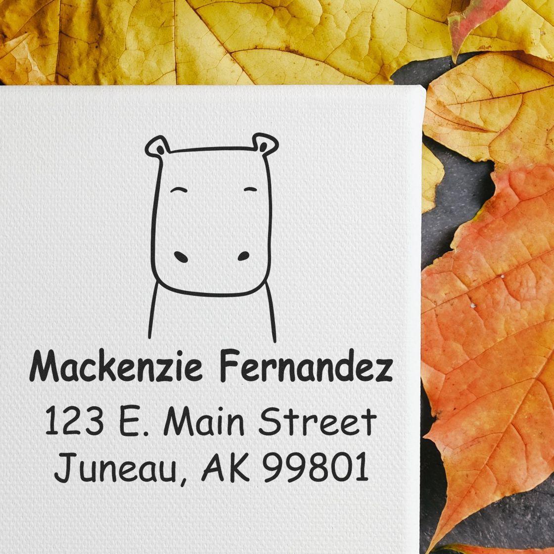 Hopeful Hippo Pre-Inked Handcrafted Mailing Stamper on a canvas with the text "Mackenzie Fernandez, 123 E. Main Street, Juneau, AK 99801" surrounded by autumn leaves. Personalized New Address Stamper design features a simple hippo illustration.
