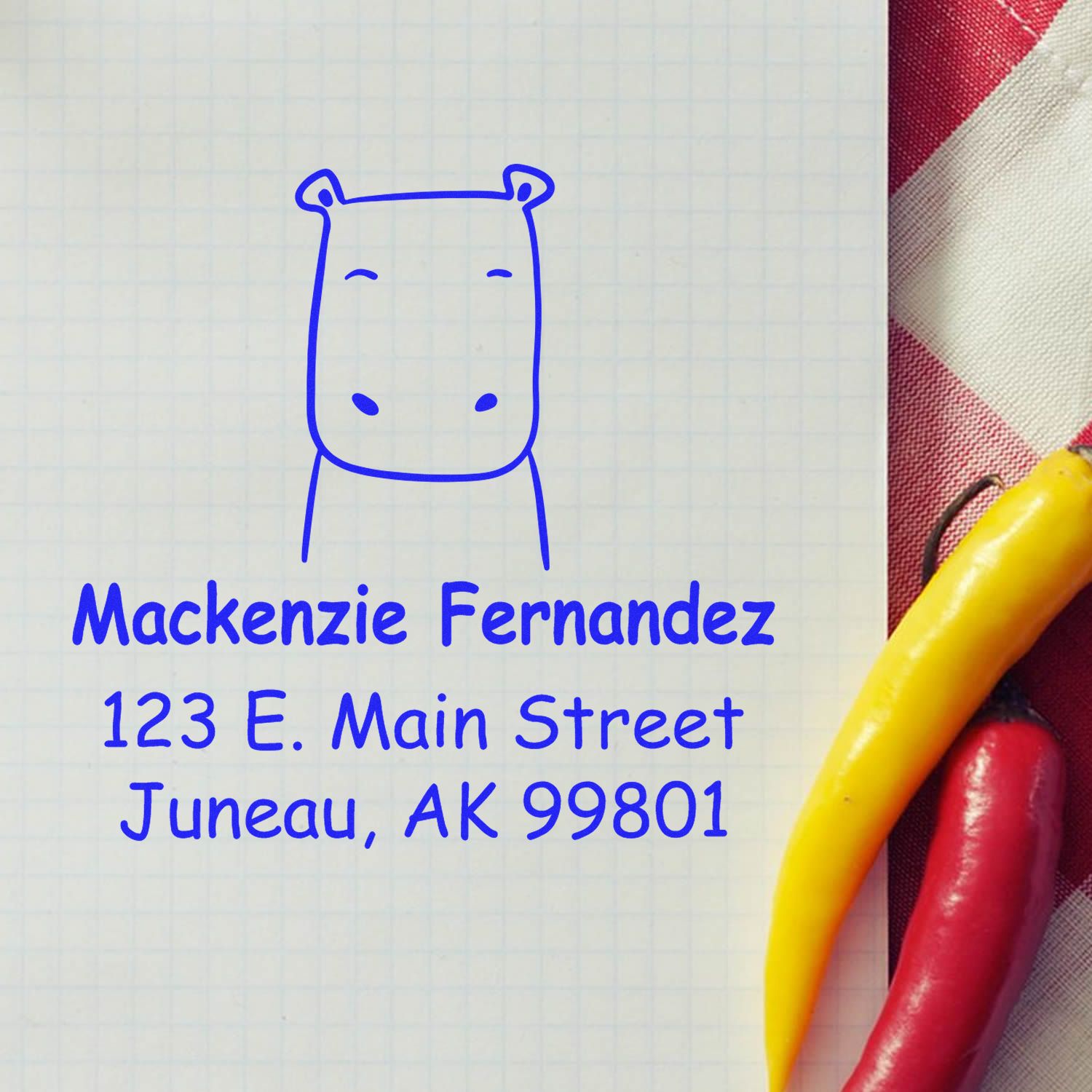 Self-Inking Cartoon Animal Hopeful Hippo Personalized Address Stamp on graph paper. The Customized Address Stamp displays "Mackenzie Fernandez, 123 E. Main Street, Juneau, AK 99801" in blue ink, next to red and yellow chili peppers on a checkered cloth.