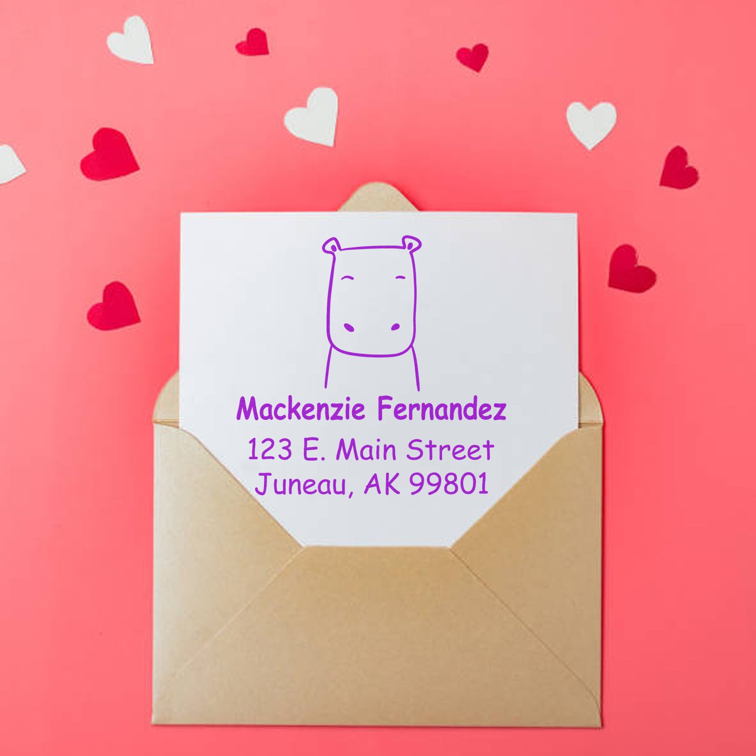 A pink background with scattered hearts features an envelope containing a card stamped with a purple hippo design. The text reads "Mackenzie Fernandez, 123 E. Main Street, Juneau, AK 99801." Keywords: Hopeful Hippo Pre-Inked Handcrafted Mailing Stamper, Personalized Return Mail Stamper.